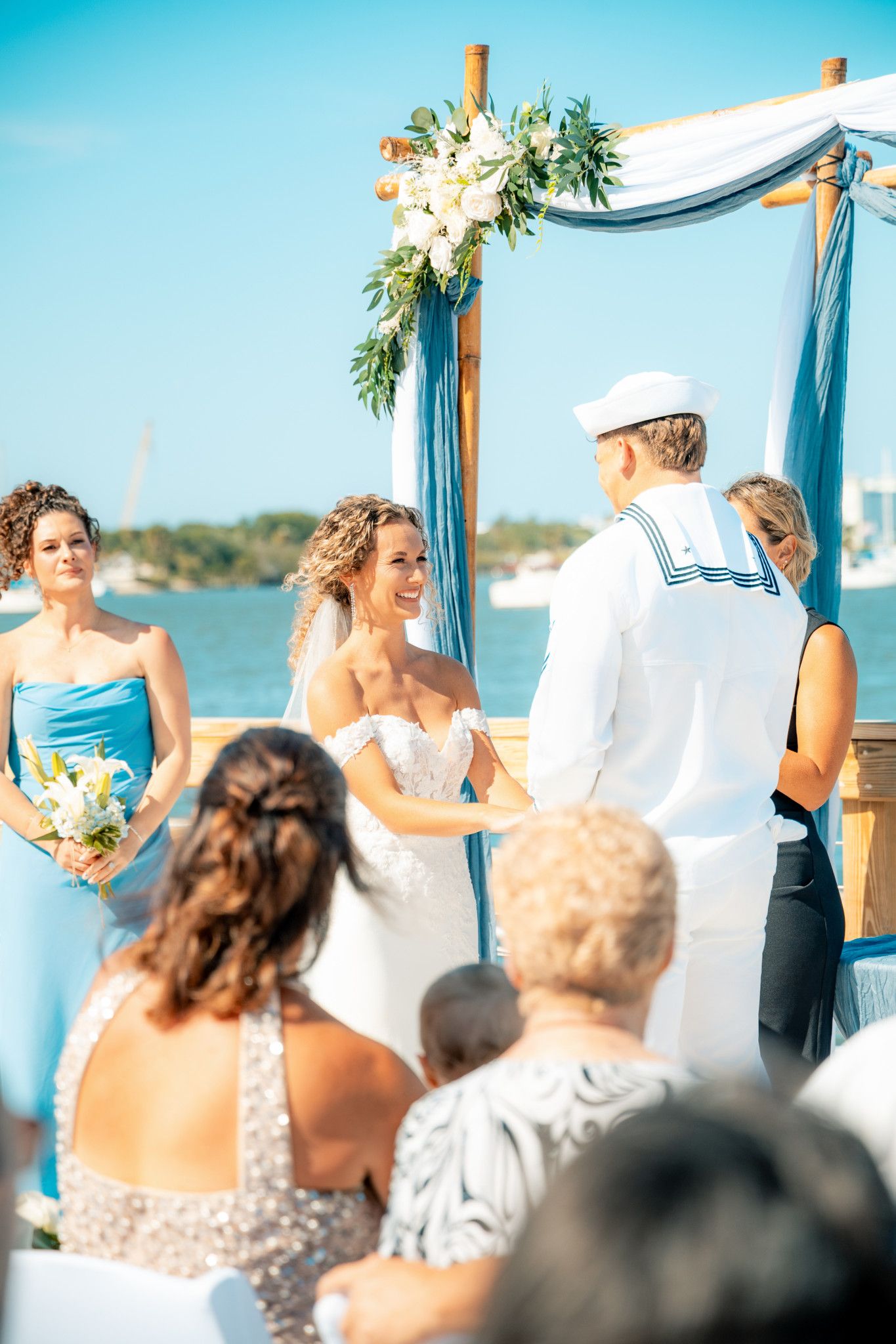 Outdoor beach wedding ceremony with bride in white gown and groom in sailor uniform under blue draped floral arch.