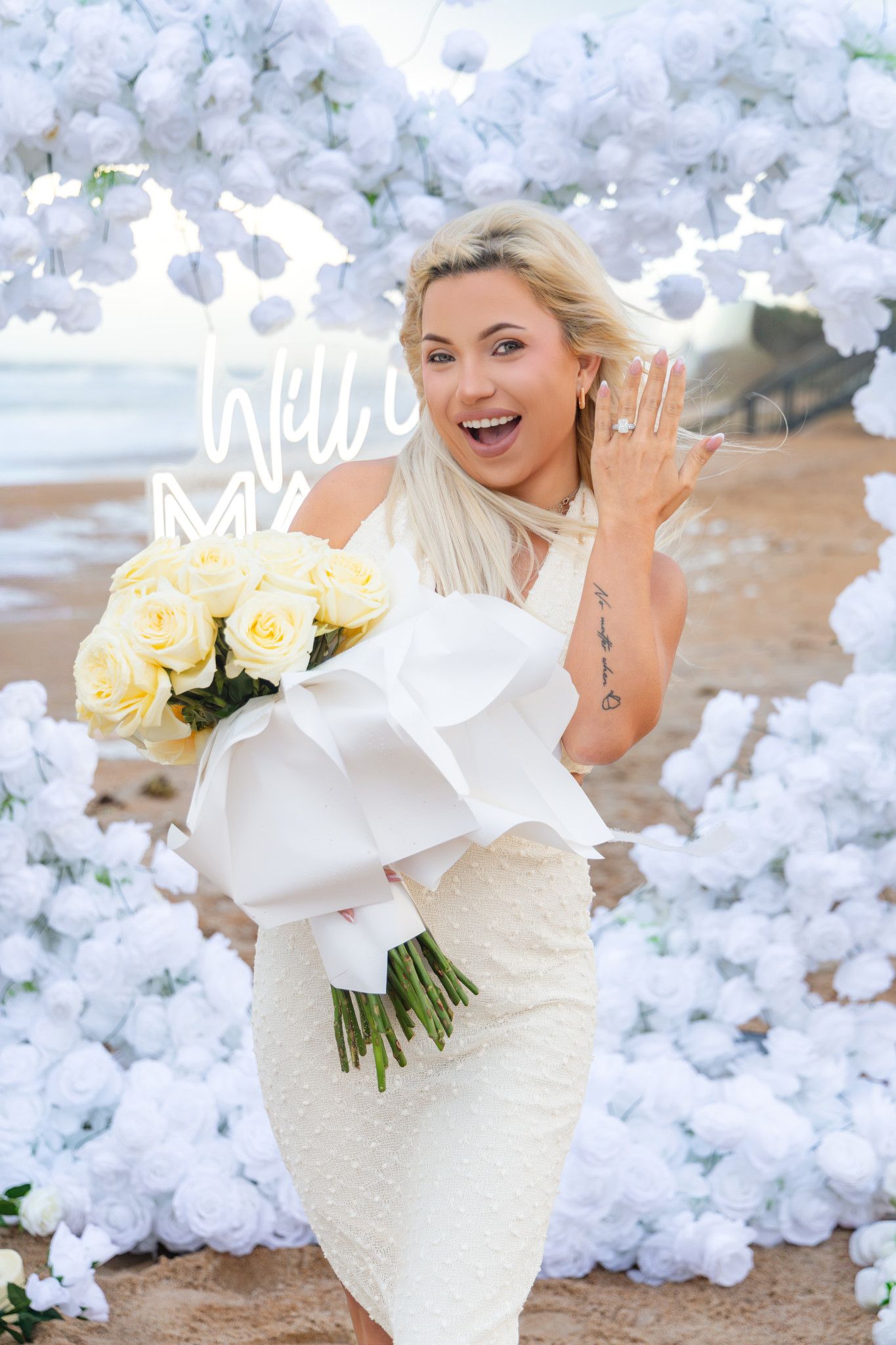 Smiling woman in a white dress holding a yellow rose bouquet and showing her ring beside a white flower arch on a beach proposal.