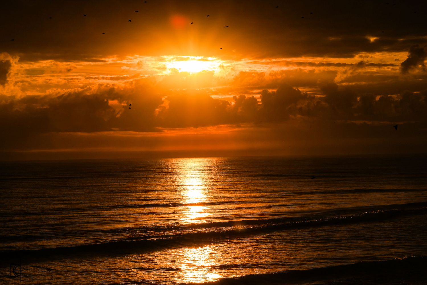 Dramatic orange sunset over calm ocean waters with sunlight breaking through clouds and reflecting on gentle waves.