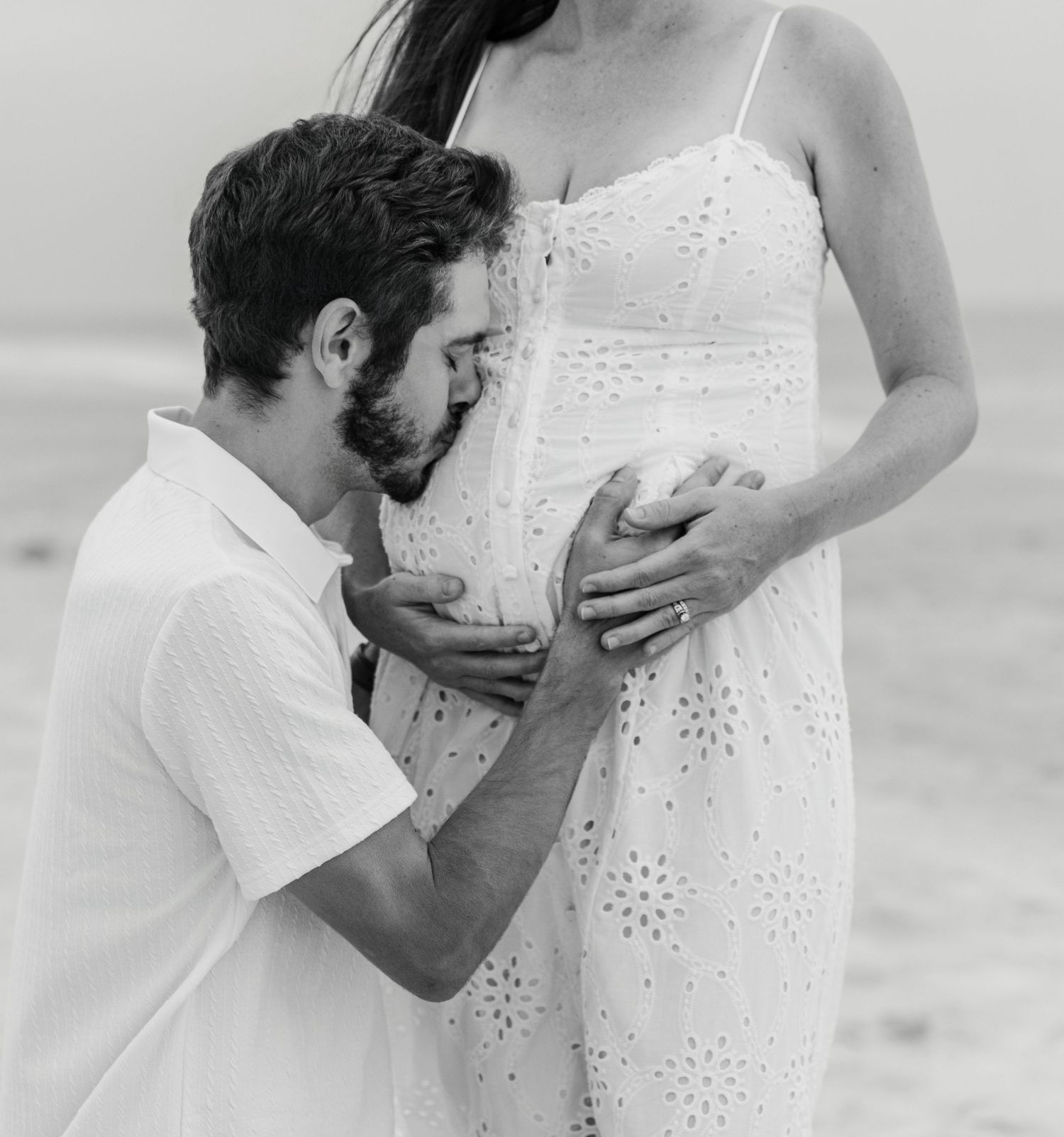 Black and white maternity photo of a man kissing pregnant woman's belly on a beach.