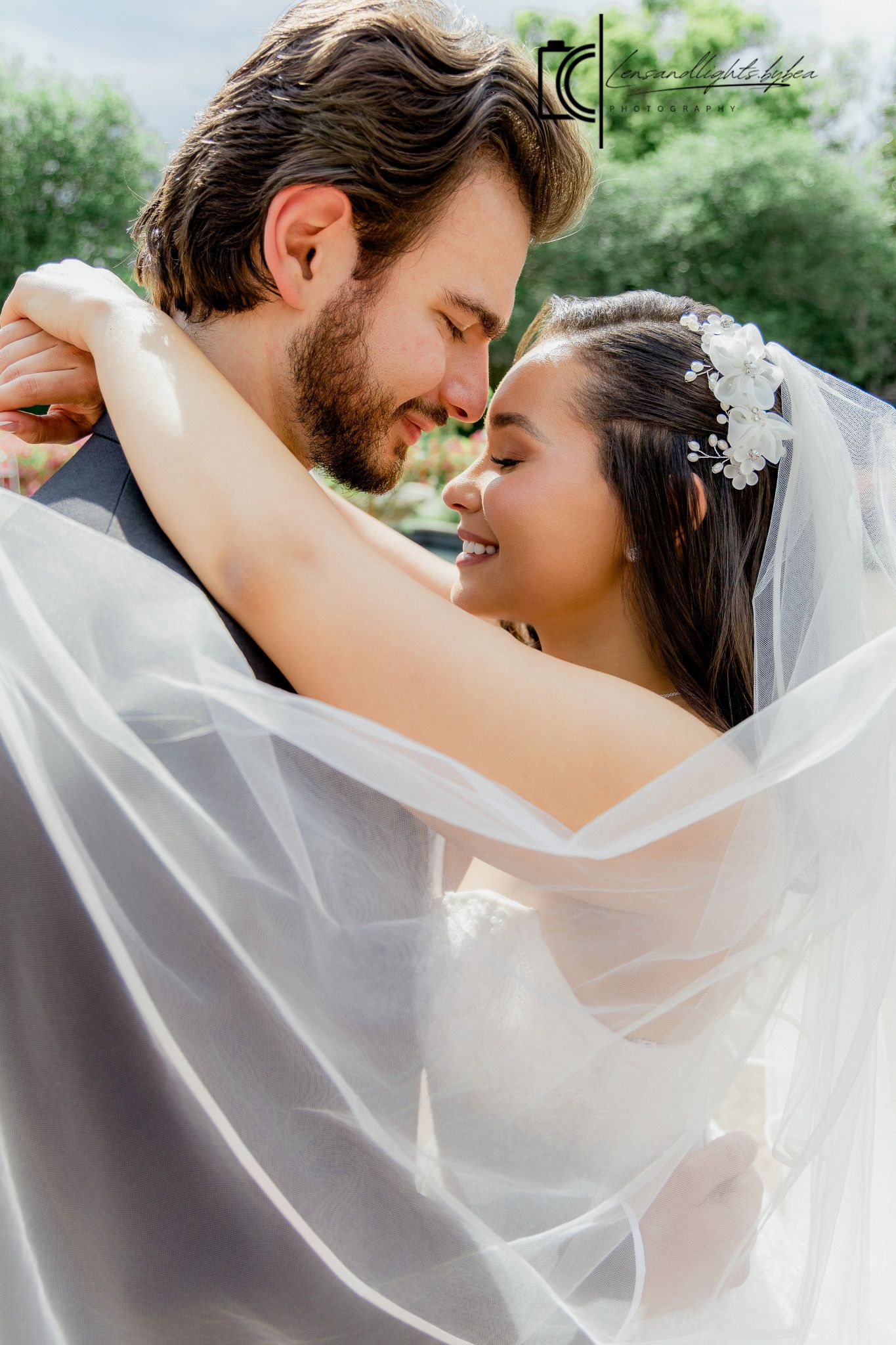 A bride and groom share a romantic moment under a flowing white veil, smiling at each other outdoors.