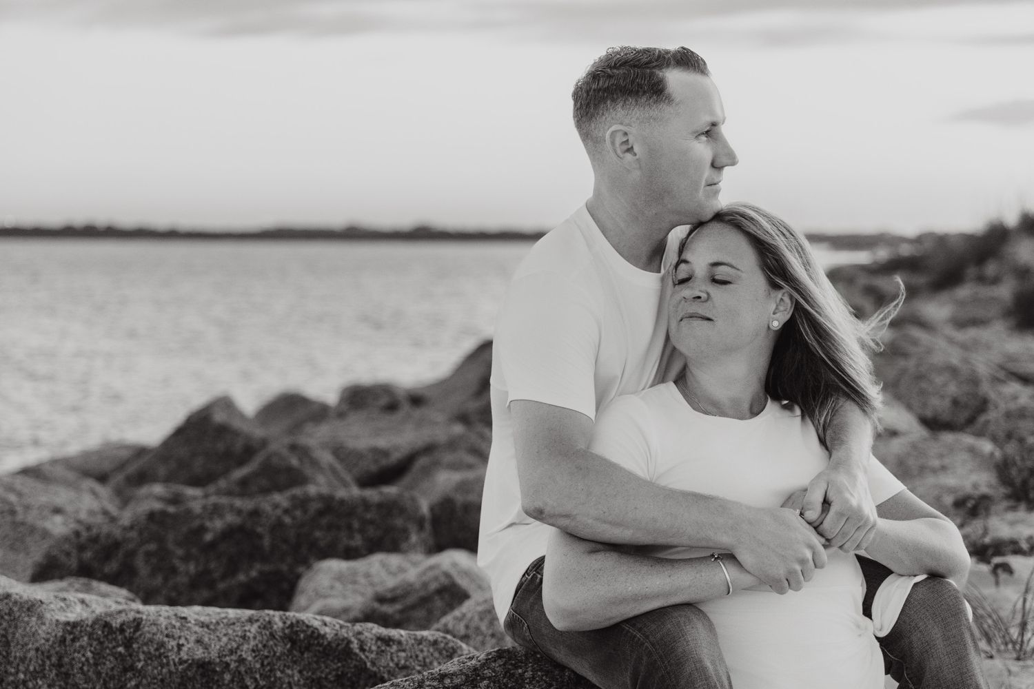 Couple embracing on rocky shoreline at sunset in black and white, romantic engagement photo by the water.