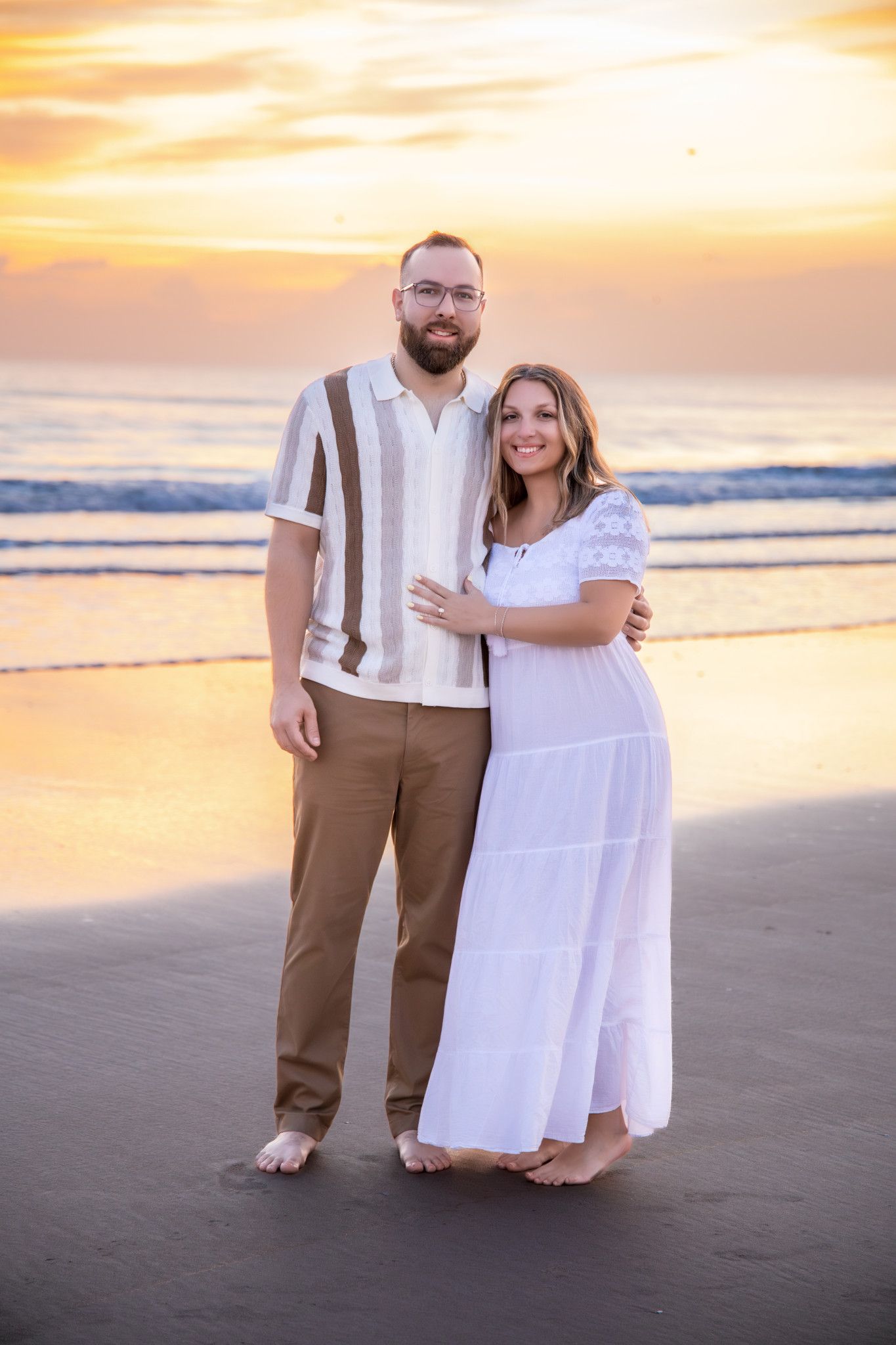 A couple stands together on a sandy beach at sunset wearing white and brown attire for a romantic photoshoot.