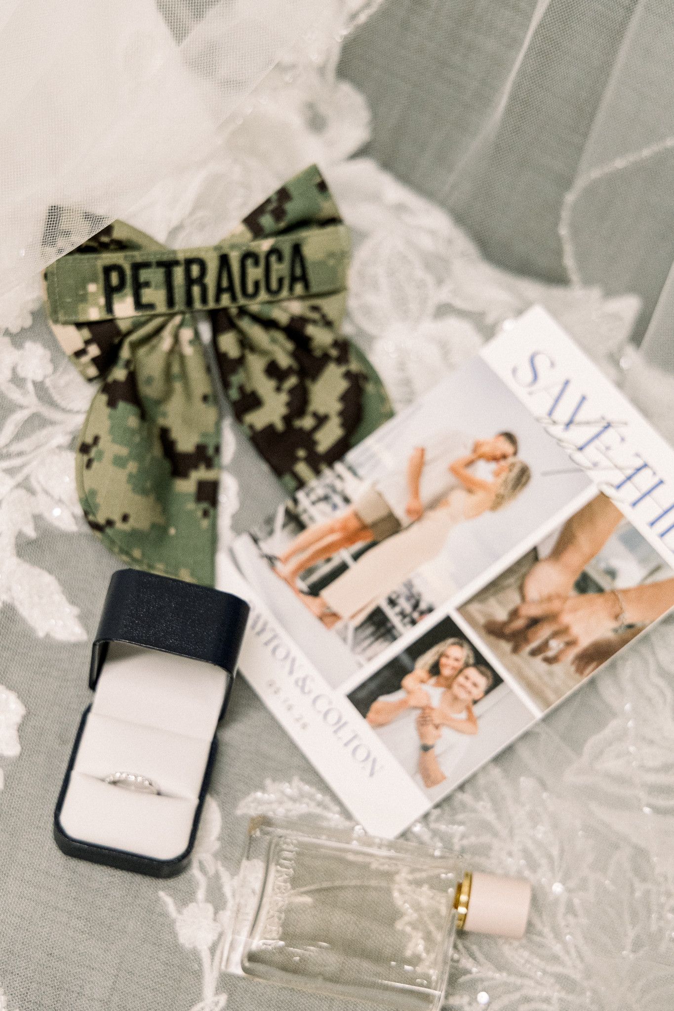 Military wedding flat lay with camouflage bow, diamond ring in box, save the date cards, and perfume bottle on lace veil.