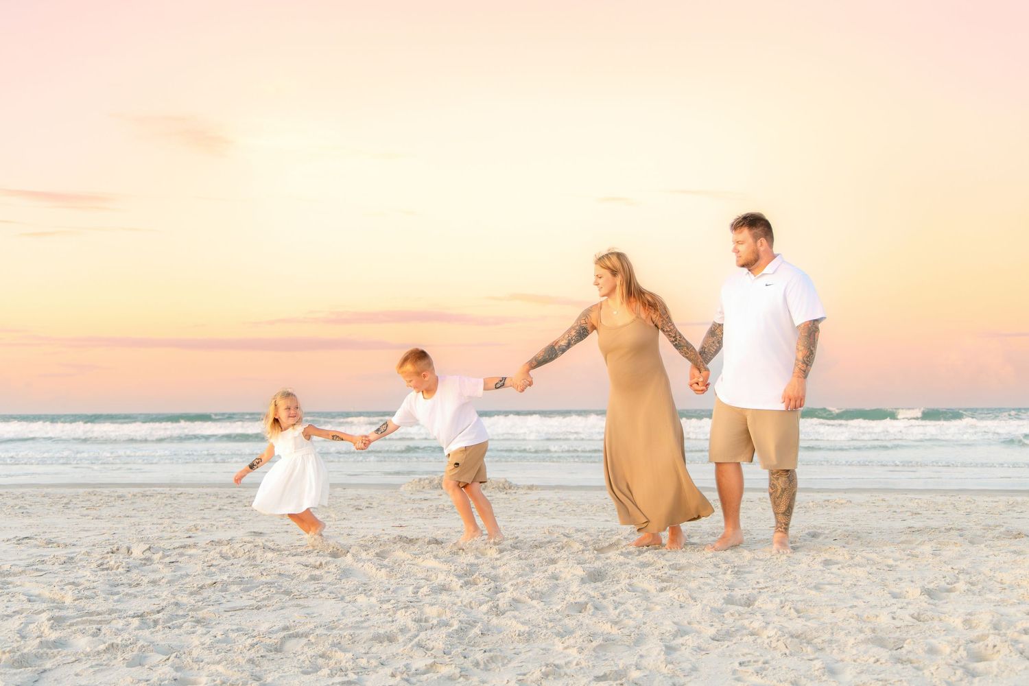 Family of four holding hands walking on a sandy beach at sunset with pastel pink and orange sky.