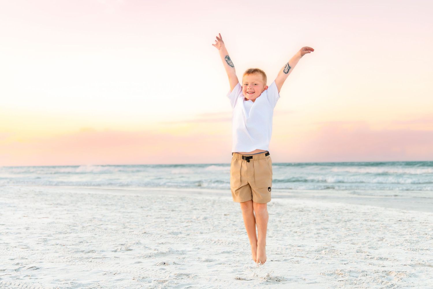 Joyful boy jumping with arms raised on a sandy beach at sunset, wearing white shirt and khaki shorts.