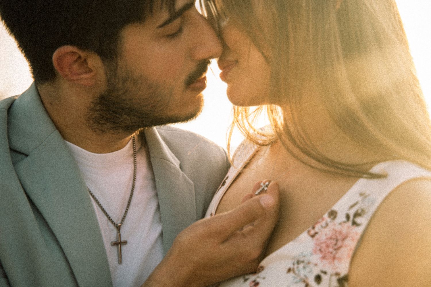 Romantic couple sharing an intimate moment in golden sunlight, man in blue blazer with cross necklace leaning toward blonde woman.