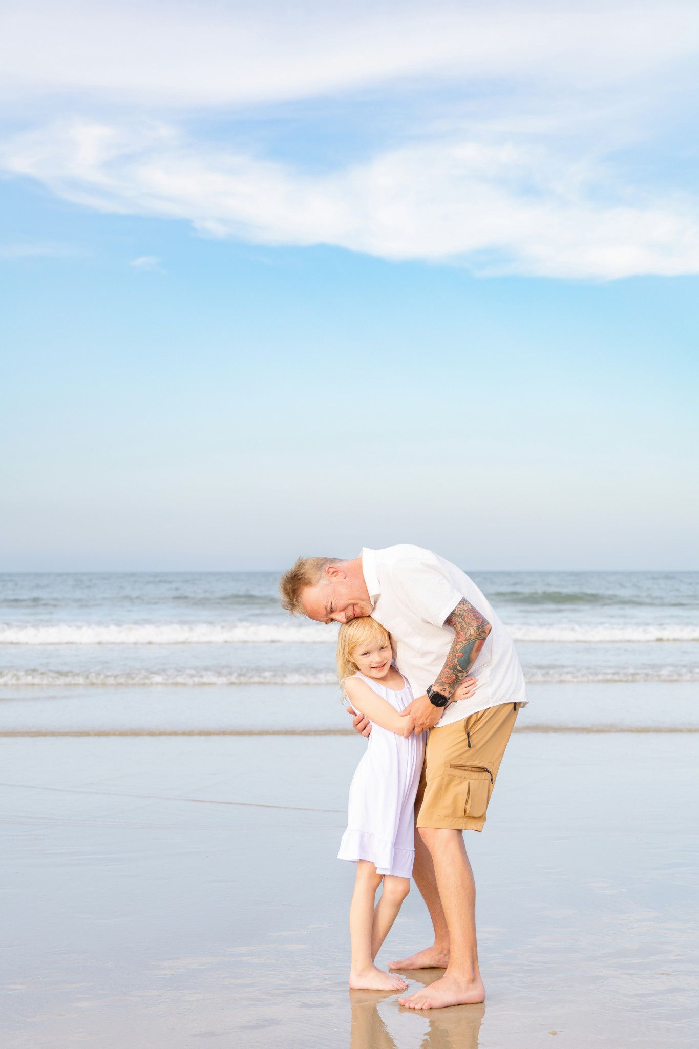 A man bending down to hug a smiling little girl on a sunny beach with ocean waves in the background.