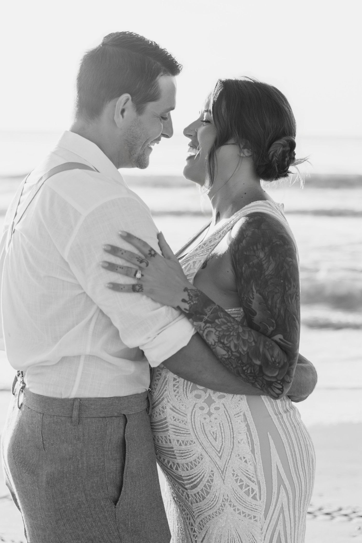 Black and white portrait of laughing bride and groom embracing on beach at sunset.
