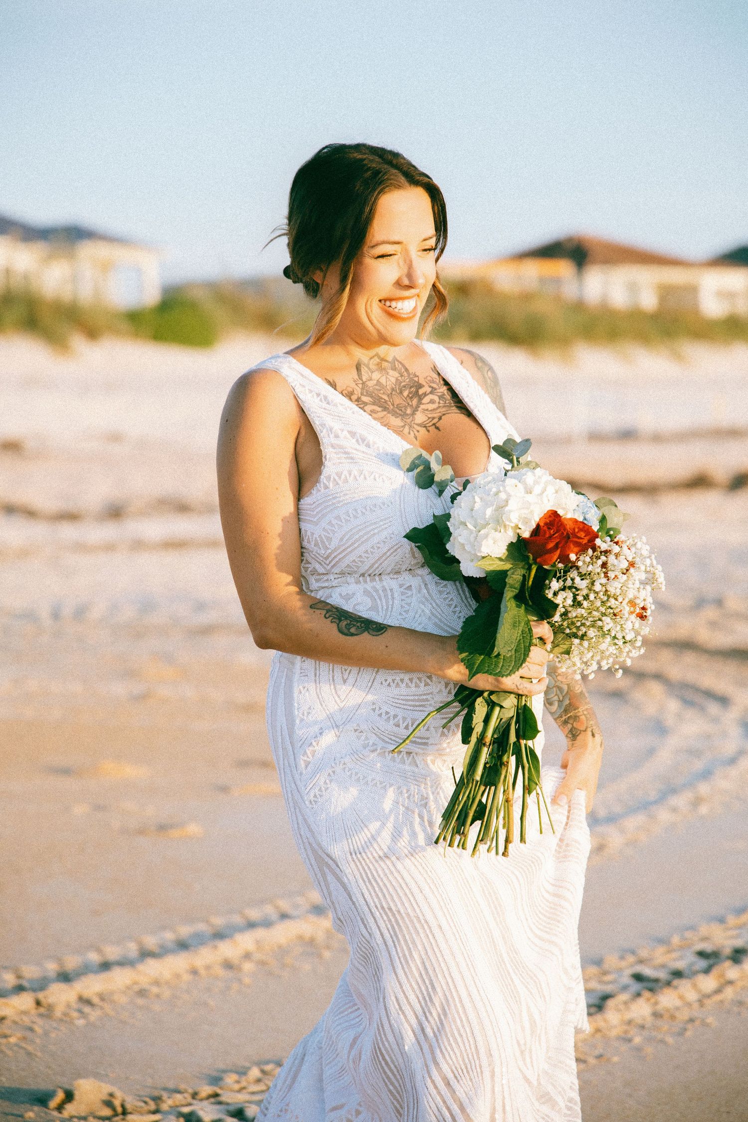 Radiant tattooed bride in lace gown laughs while holding bouquet on sandy beach.