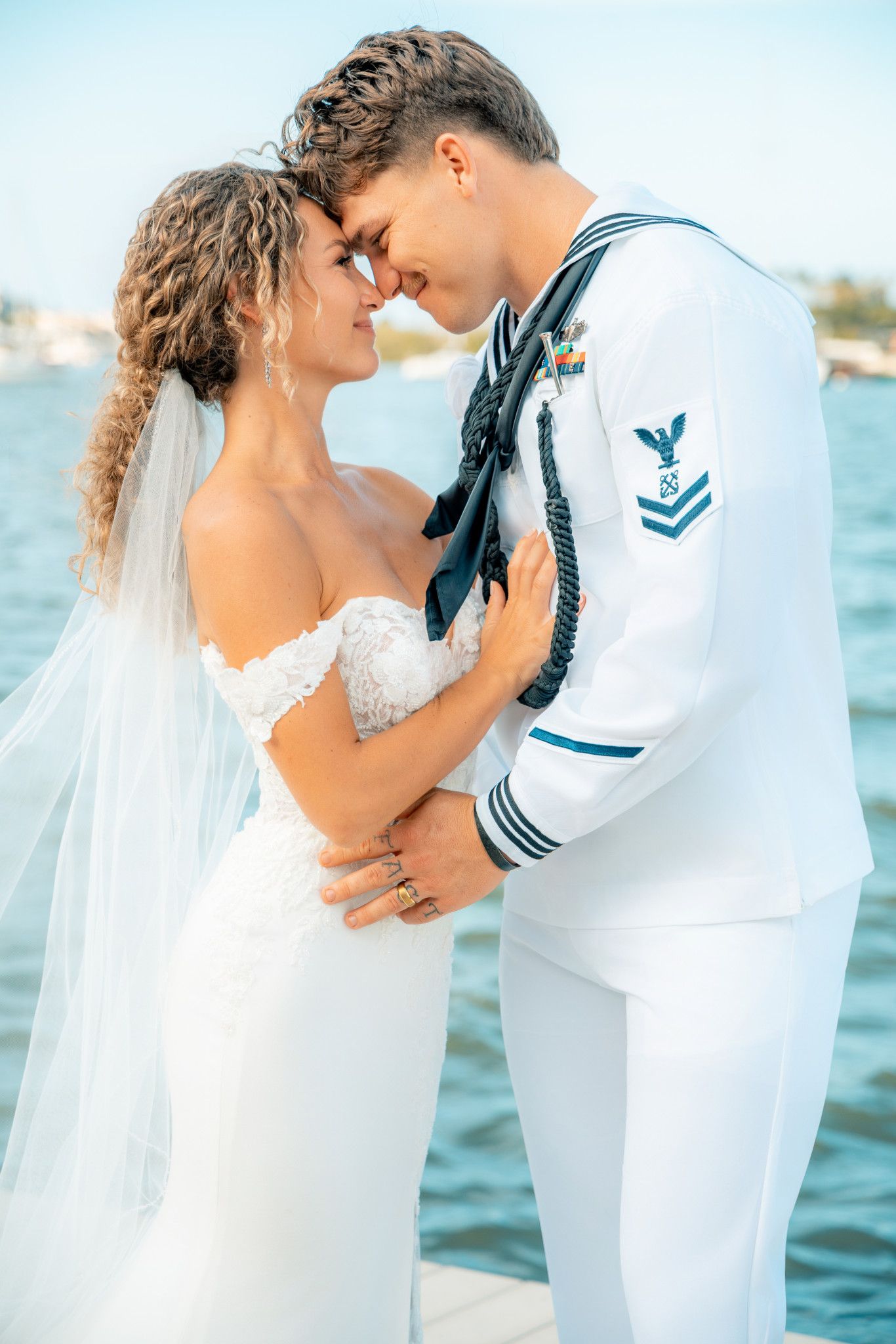 A bride and Navy sailor groom share a romantic moment dockside, touching foreheads in wedding attire near the water.