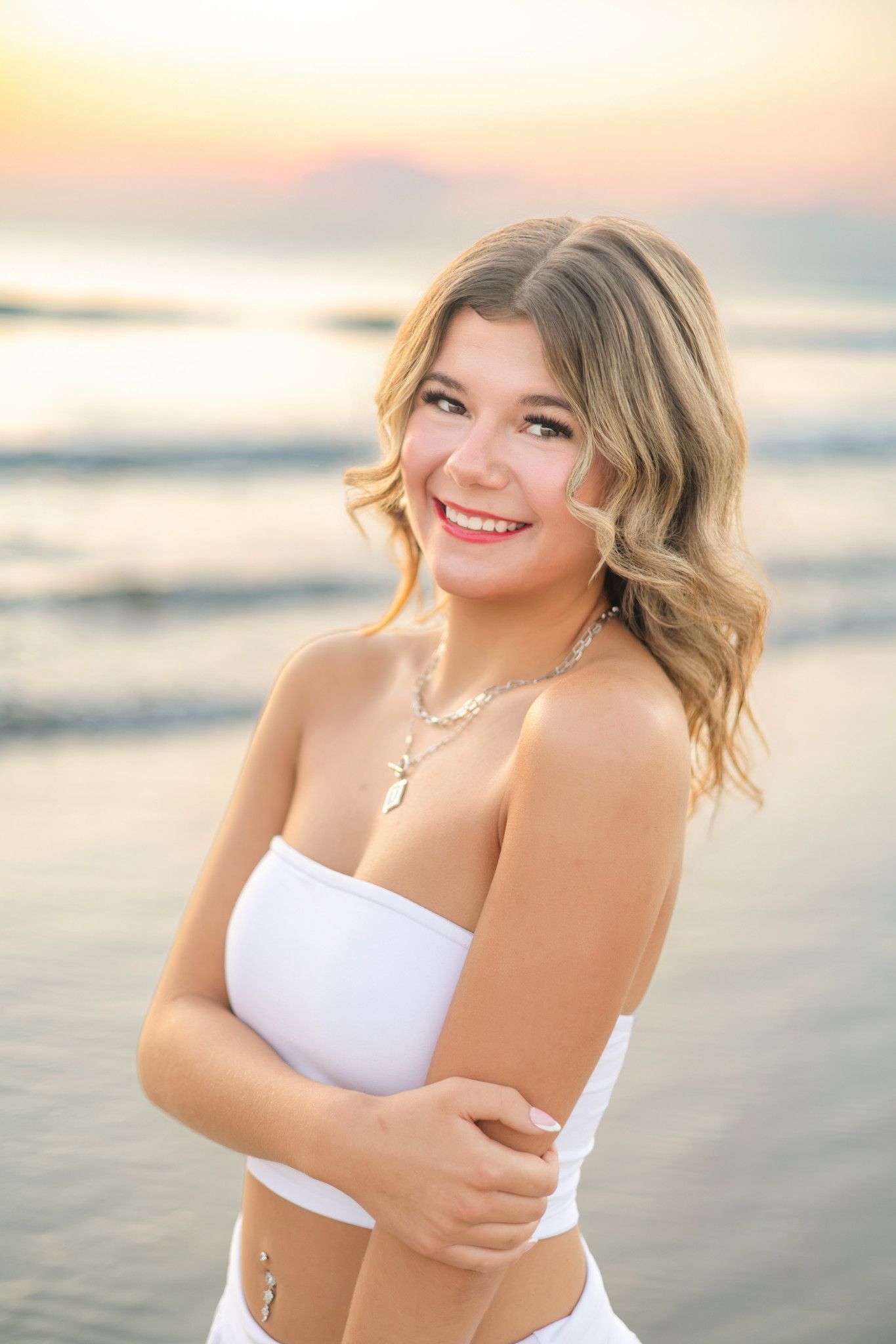 Smiling young woman in white bandeau top poses at beach during golden sunset.