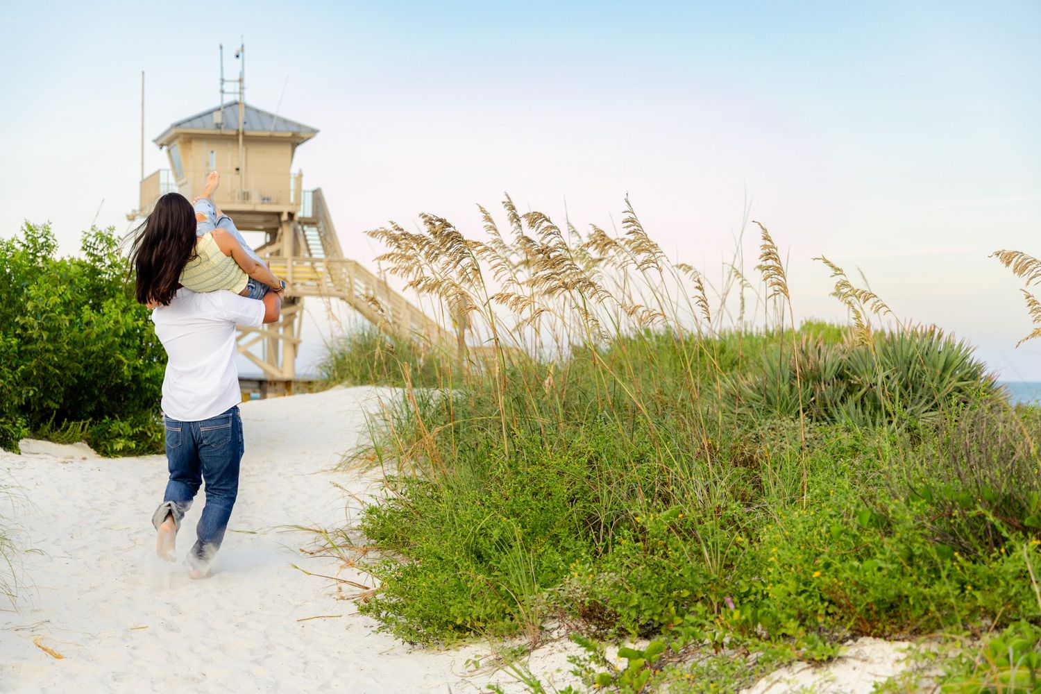 Woman walking on a sandy beach path toward a lifeguard tower surrounded by sea oats and greenery.