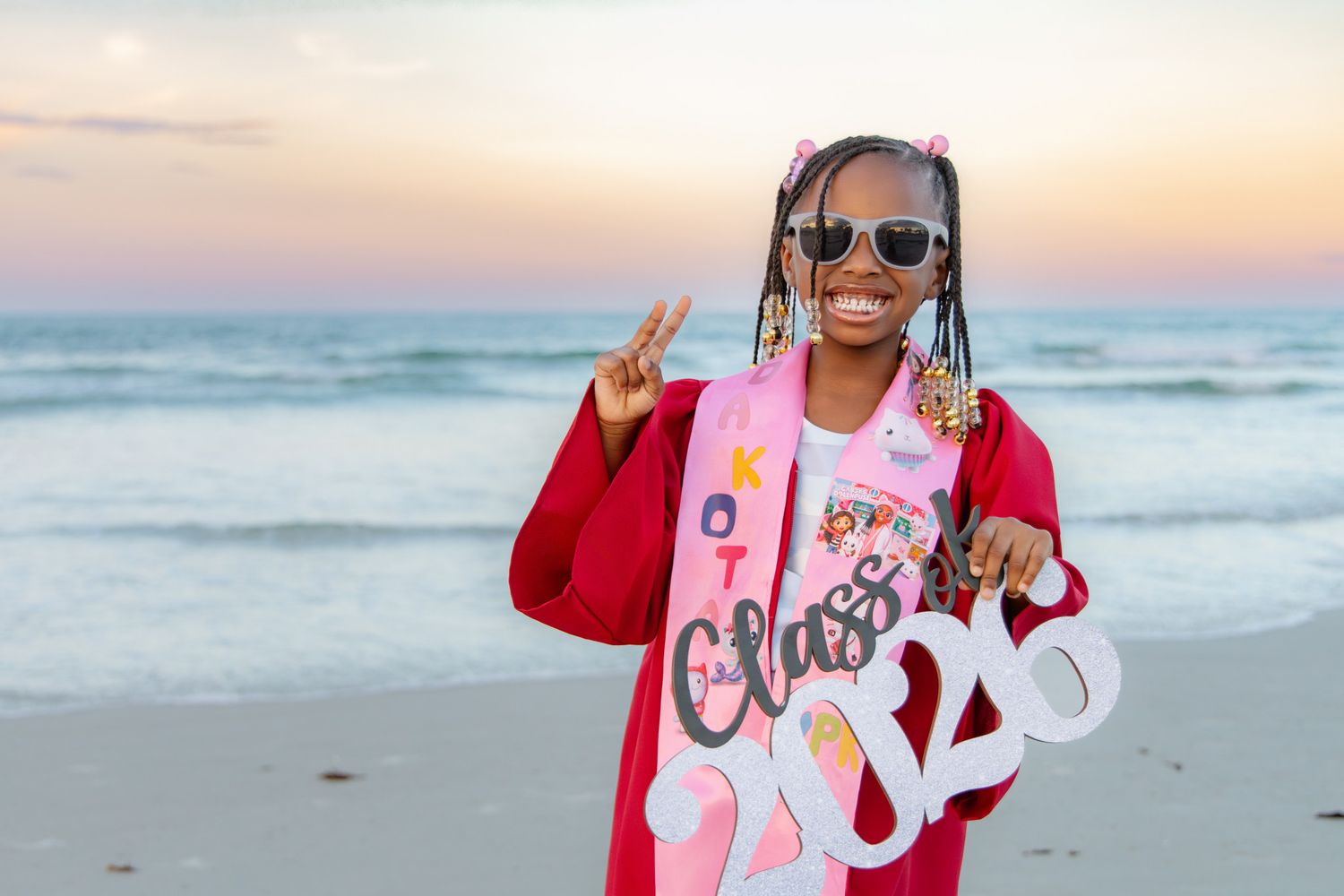 Smiling graduate in red gown holds Class of 2024 sign on beach at sunset.