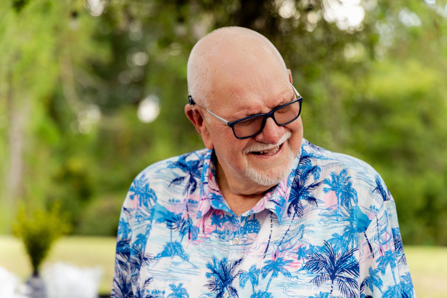 Smiling bald man with glasses wearing a colorful tropical Hawaiian shirt outdoors.