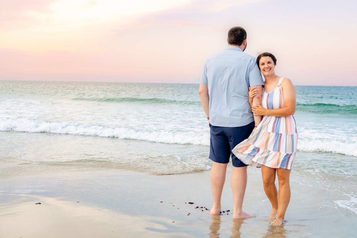 Couple standing on a sandy beach at sunset, waves gently rolling in, woman in striped dress smiling.