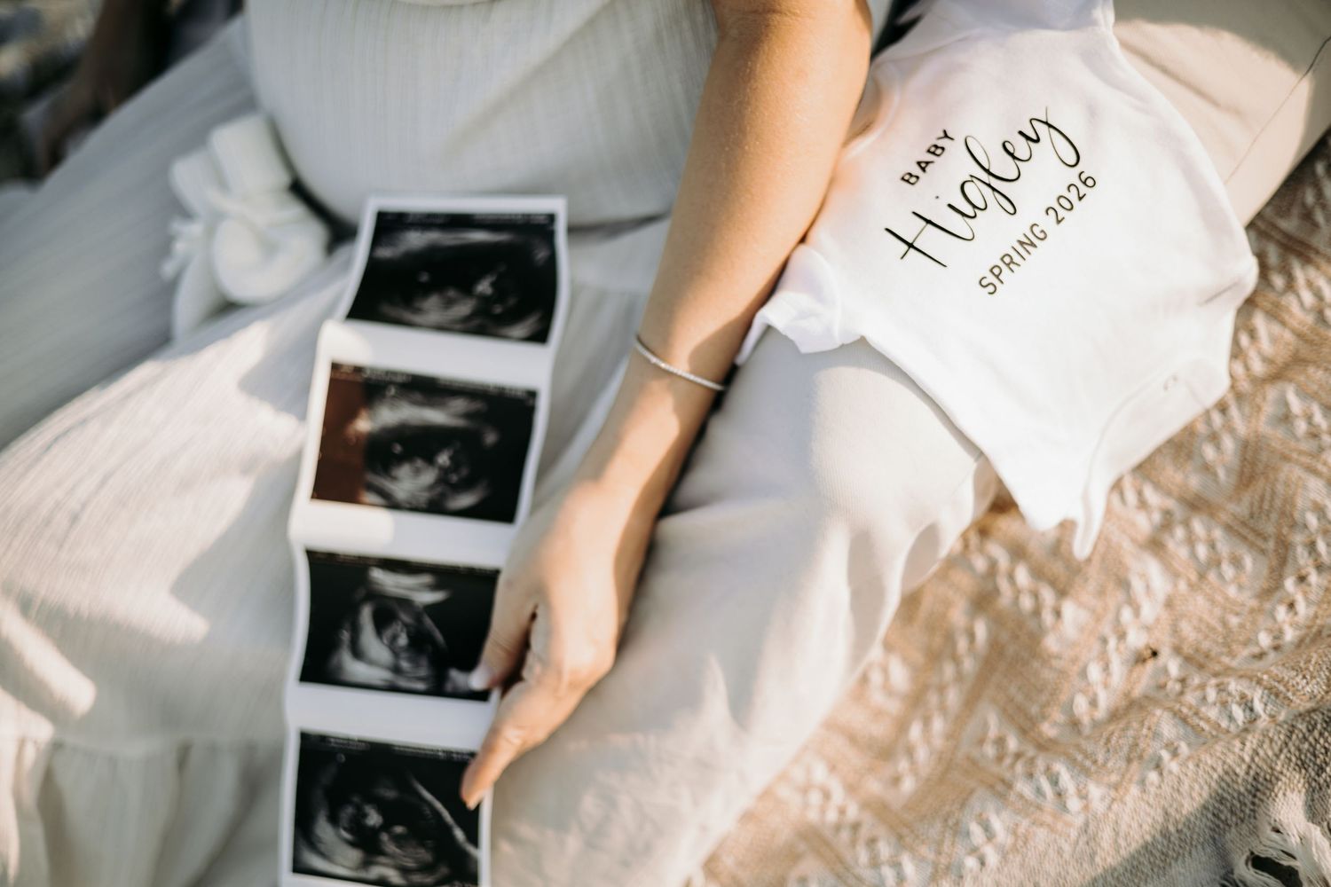 Ultrasound photos and pregnancy announcement card laid on white textured fabric with someone's hand holding the images.