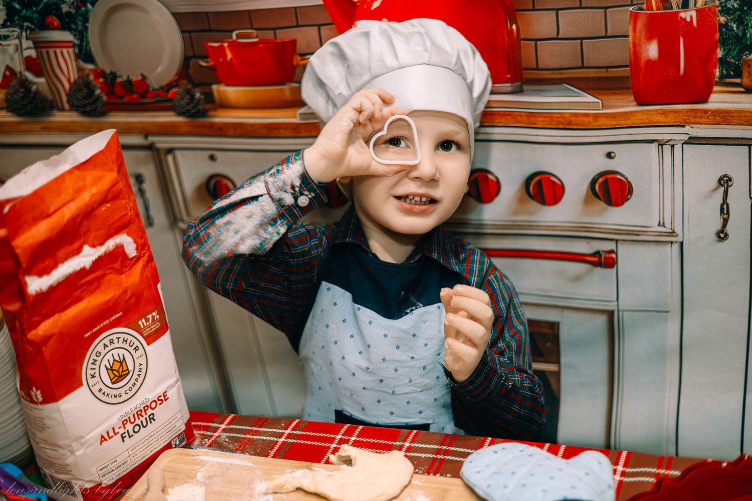 A young chef in a white hat stands at a kitchen counter with baking ingredients and red decorative elements.