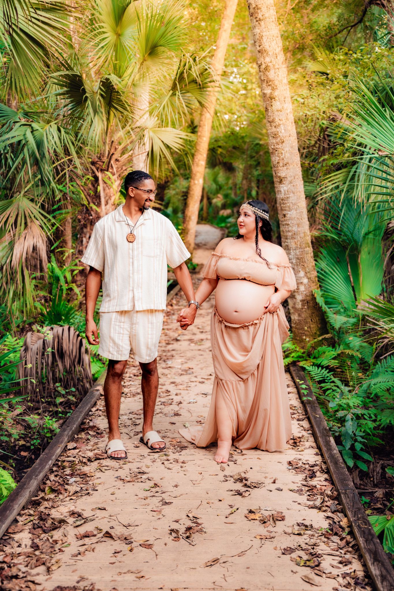 Pregnant couple holding hands walking on sandy path surrounded by lush tropical palm trees during maternity photoshoot.