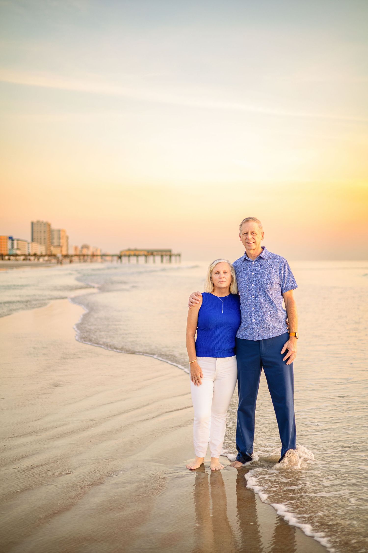 Senior couple standing barefoot on a sandy beach at sunset, wearing blue outfits with a pier and skyline in the background.