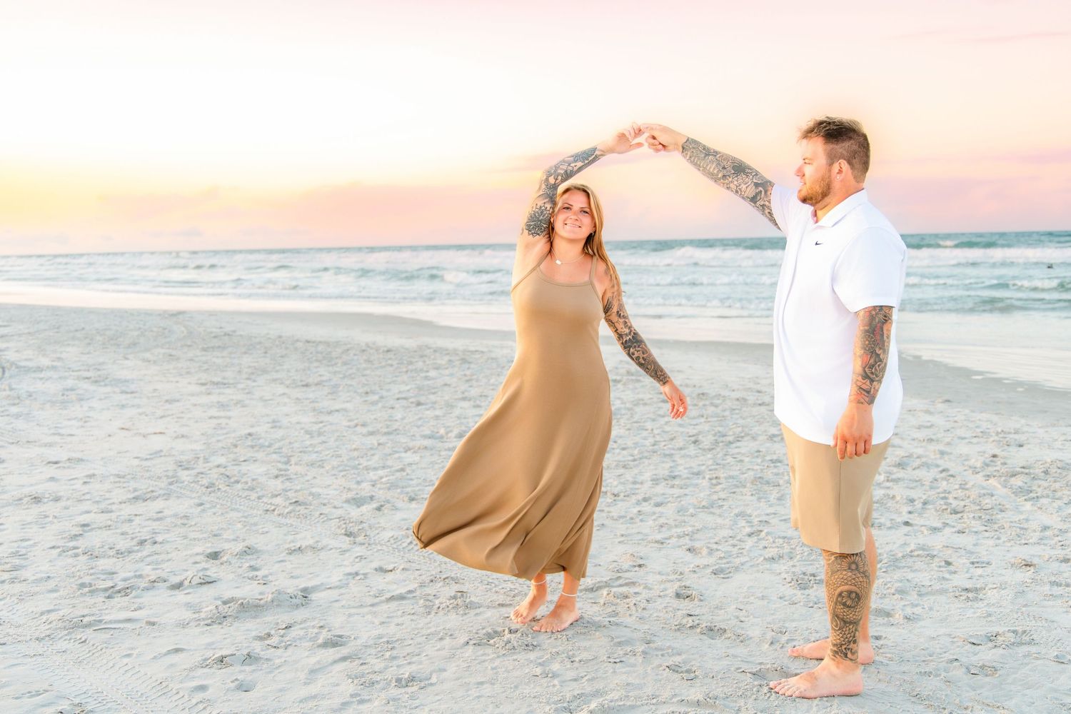 Couple dancing on a sandy beach at sunset, woman in flowing tan dress, man in white shirt, both with tattoos.