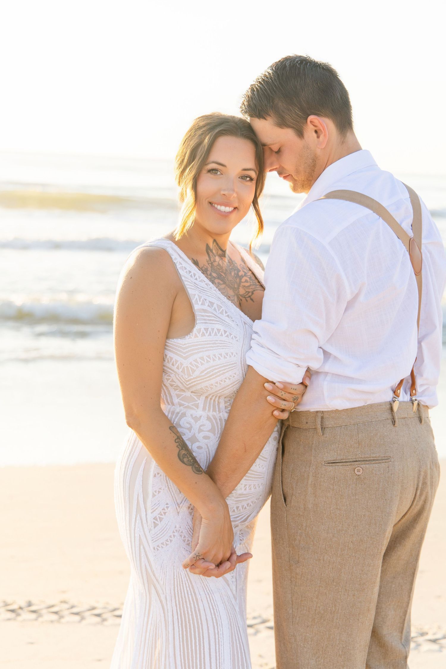 Bride and groom holding hands on beach at golden hour, groom wearing suspenders and white shirt.