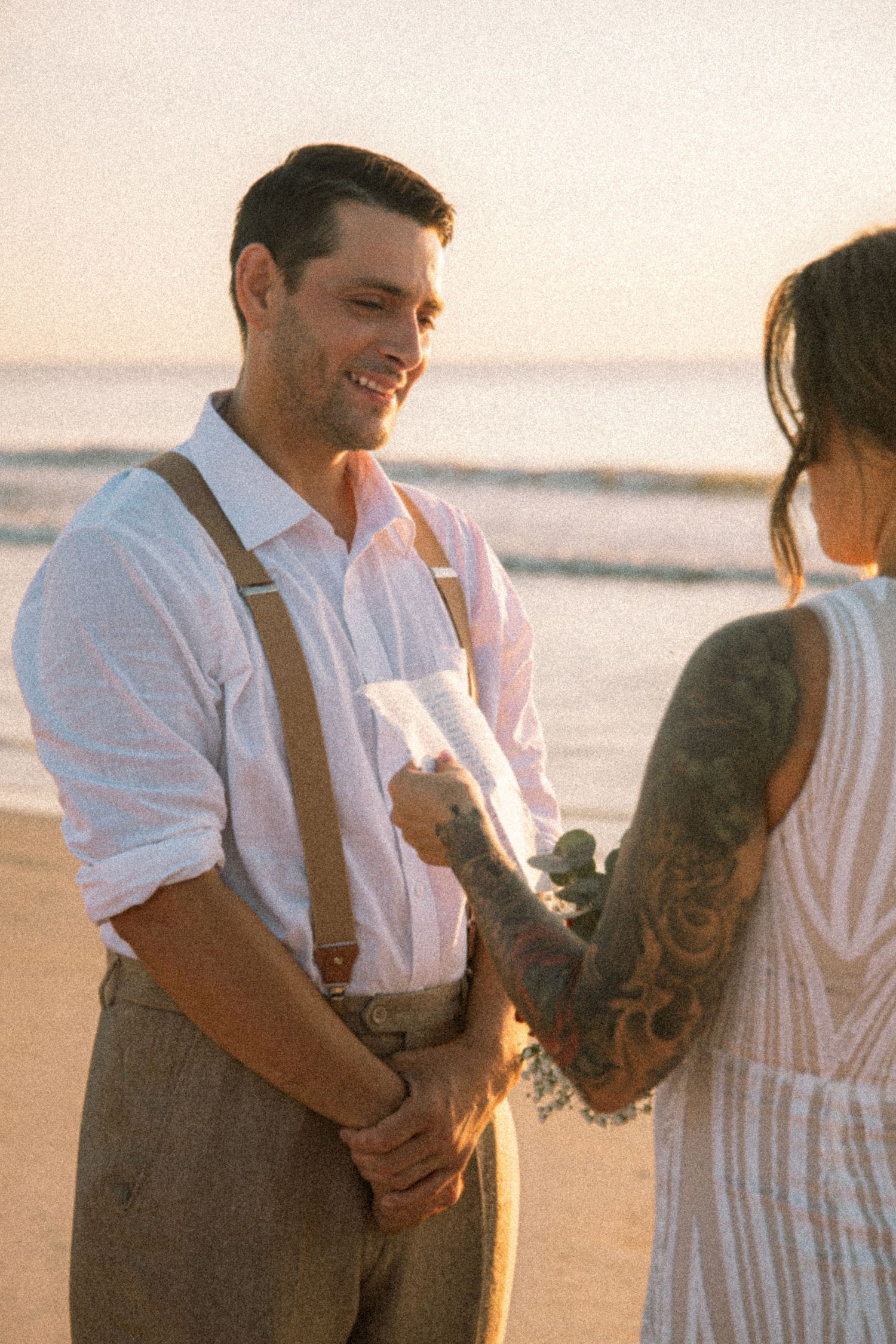 Couple exchanging vows on a beach at sunset, groom in suspenders smiling at tattooed bride holding bouquet.