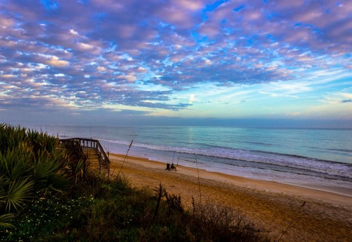Stunning beach sunset with vibrant pink and blue cloudy sky, calm turquoise ocean waves, sandy shore, and a wooden walkway.