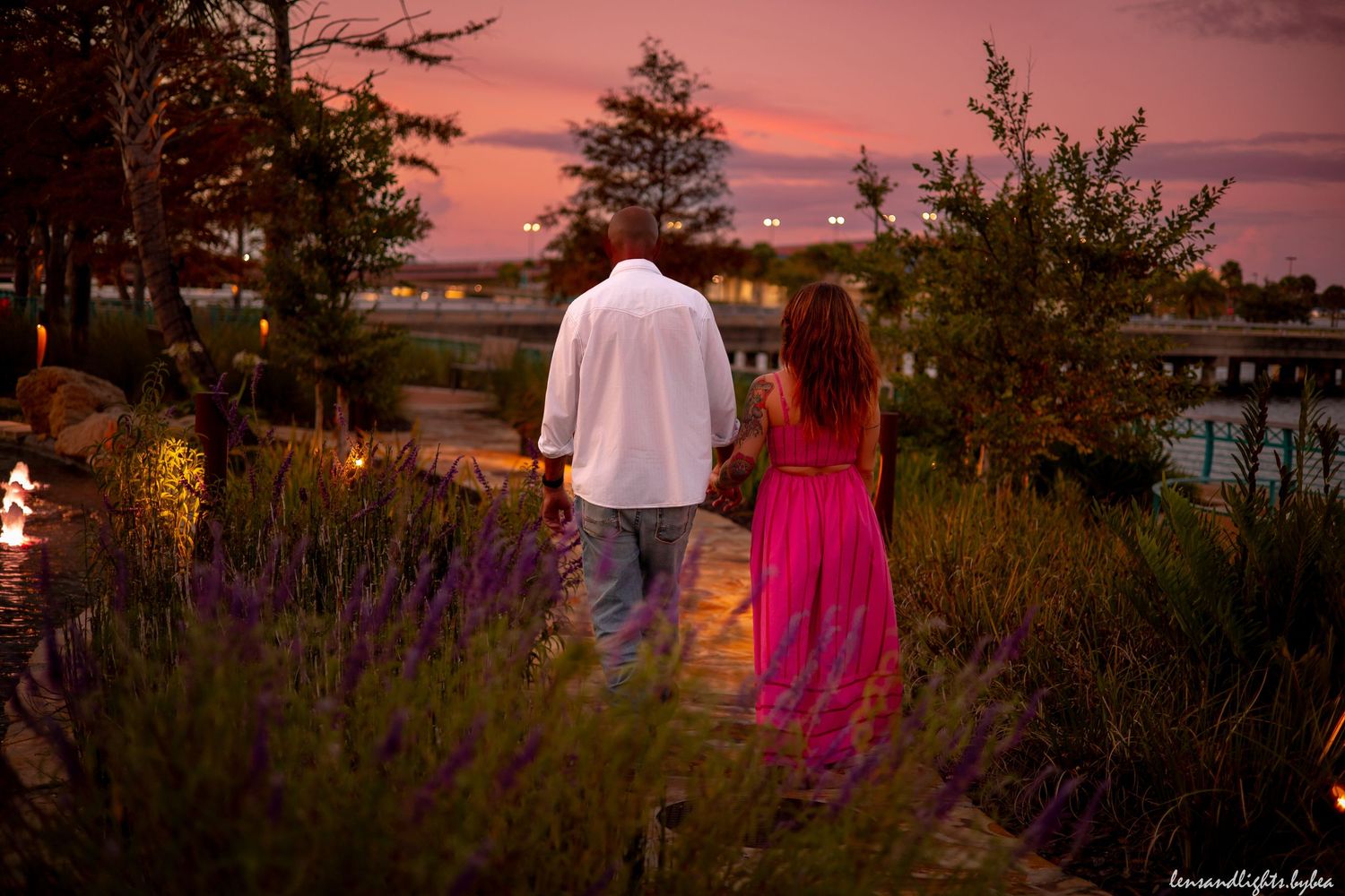 Couple walking through landscaped garden path at dusk with vibrant pink sky.
