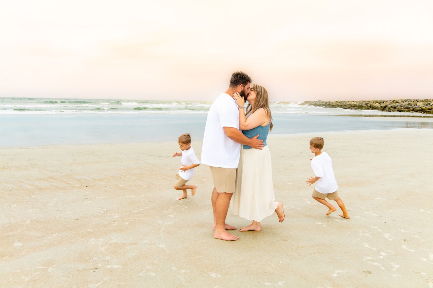 Family embracing on a sandy beach at sunset while two young children play in the background near ocean waves.
