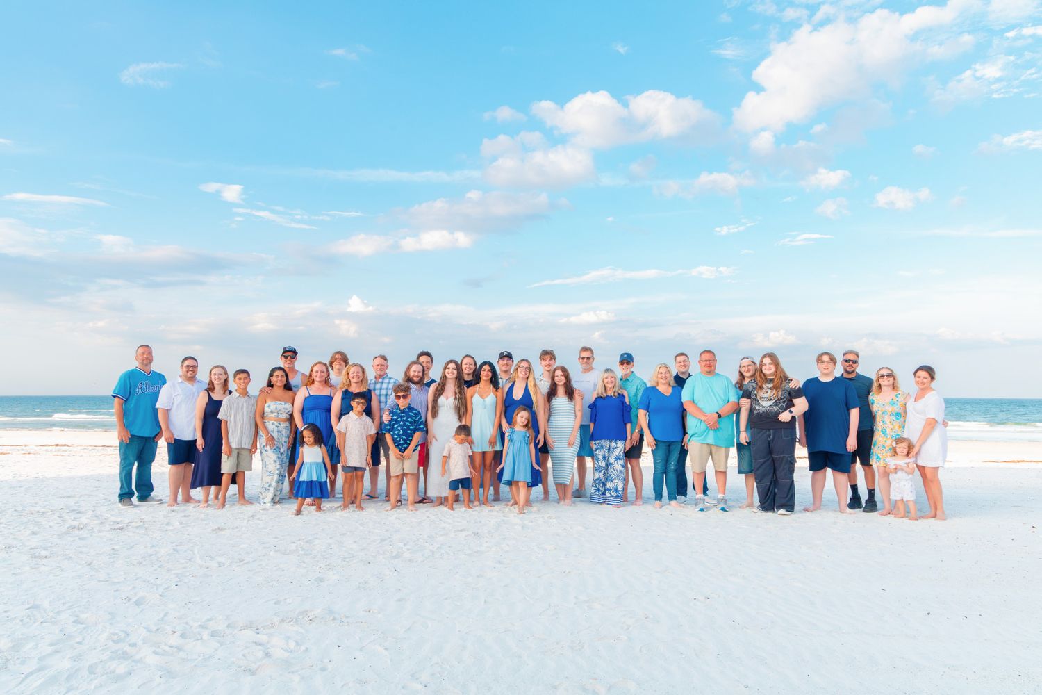 Large family group portrait on a sunny beach with white sand, blue sky, and calm ocean in the background.