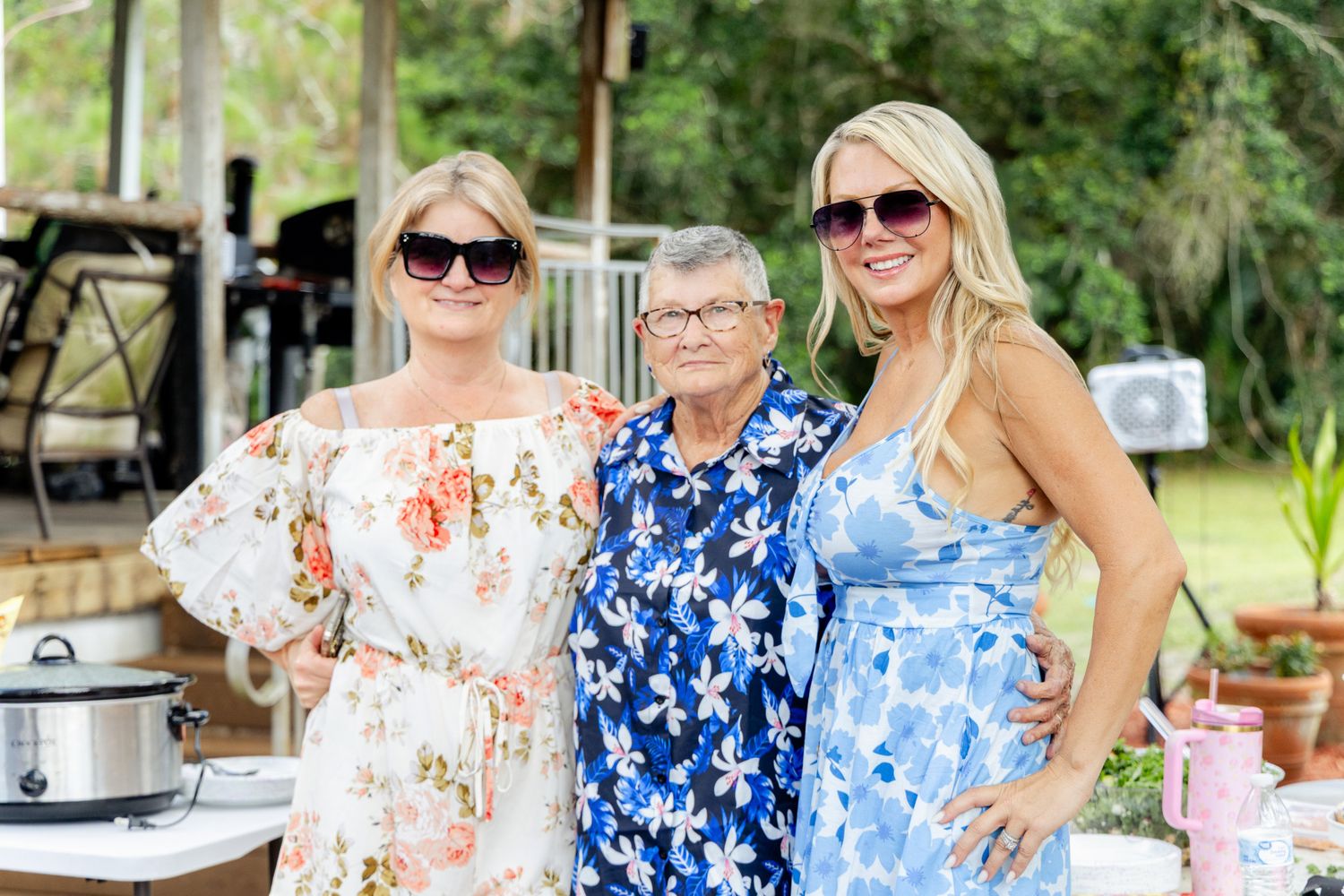 Three women wearing sunglasses and floral dresses smile together at an outdoor gathering on a sunny day.