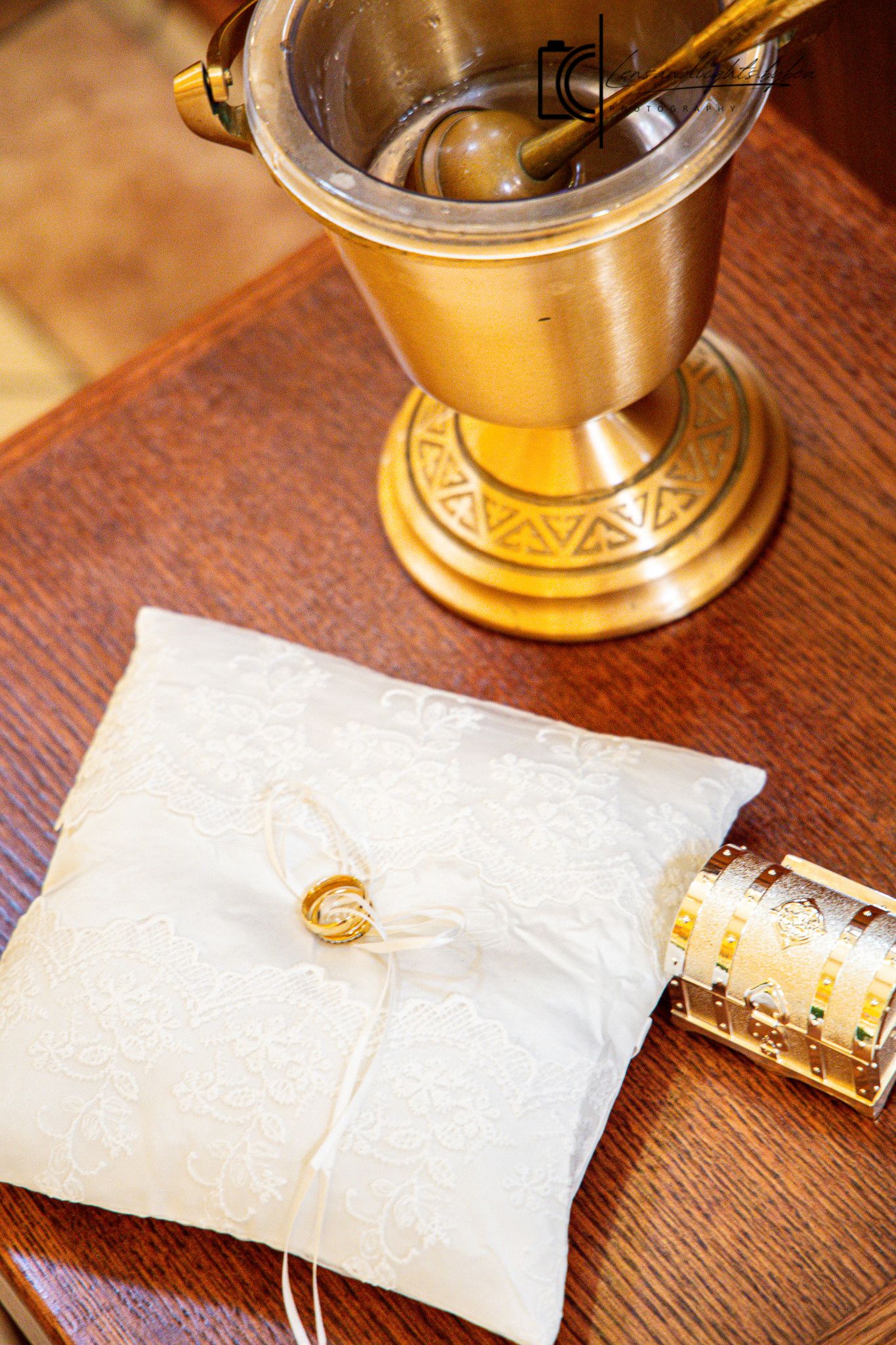 Gold wedding rings on white lace pillow beside ornate brass holy water vessel on wooden church table.