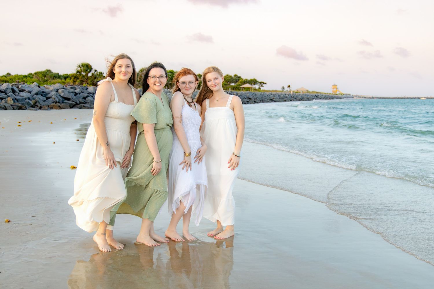 Four women in flowing dresses smiling together on a tropical beach at sunset.