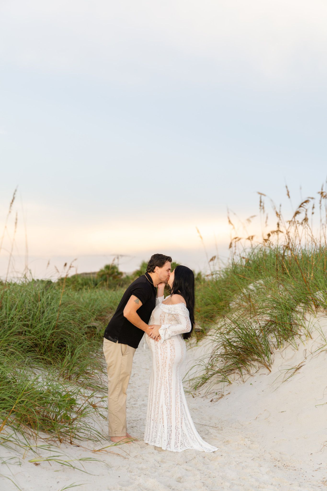 Couple sharing a romantic kiss on sandy beach dunes at sunset, woman in white lace maternity gown, man in black shirt.