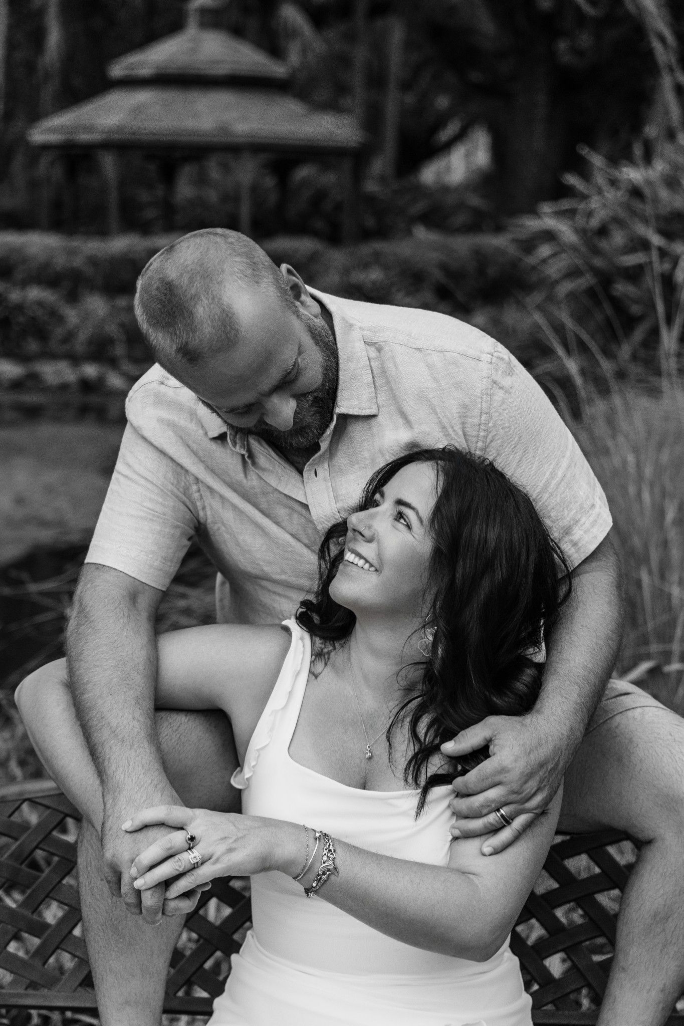 Black and white photo of a smiling couple embracing outdoors, woman seated looking up at man affectionately.