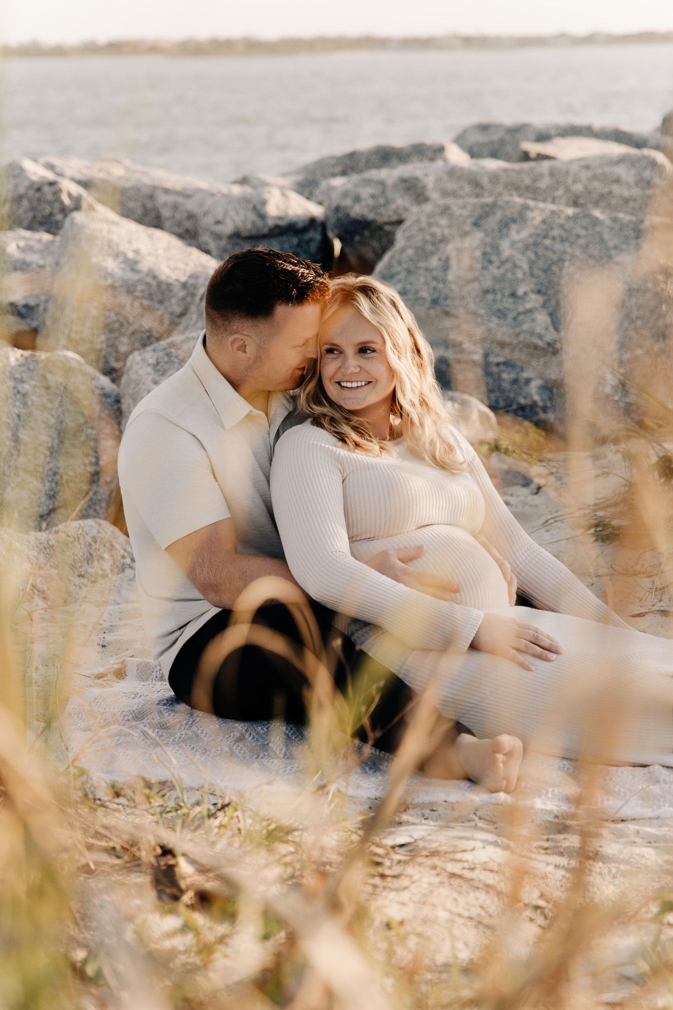 A couple shares an intimate moment on rocky coastal terrain during a maternity photoshoot at sunset.