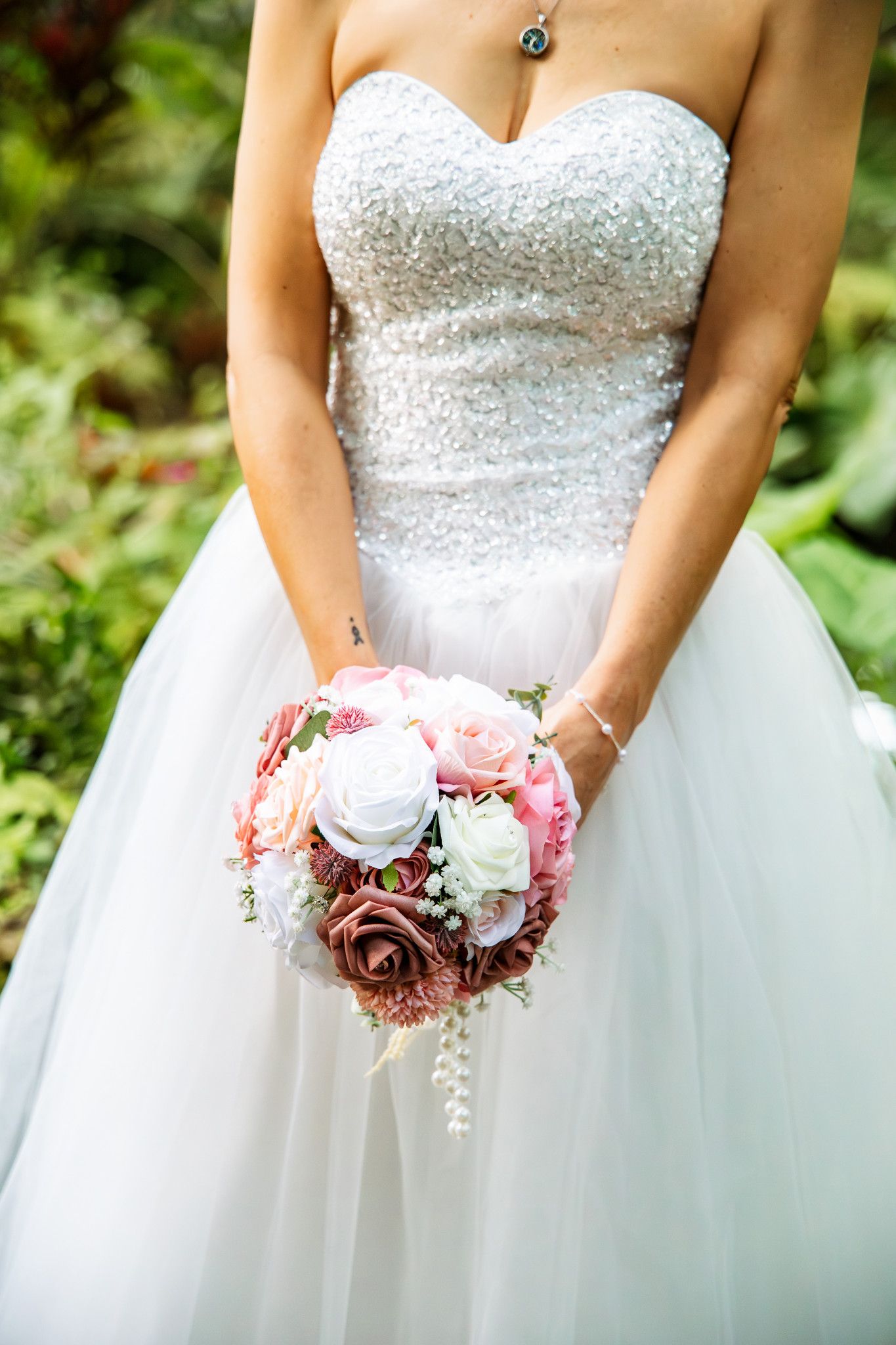 A bride in a beaded strapless wedding gown holds a romantic bouquet of pink and white roses against a garden backdrop.