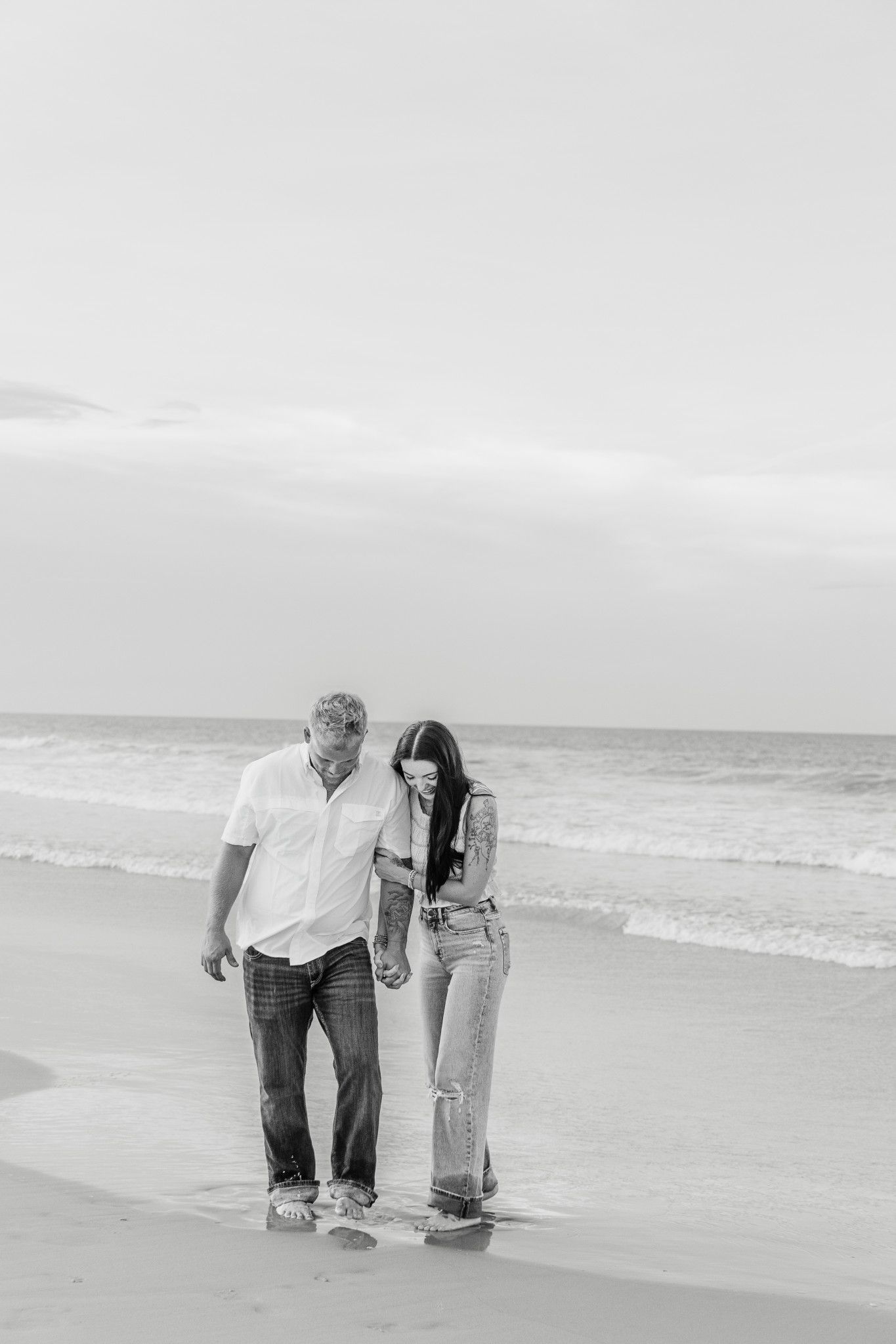 A couple holds hands walking along the shoreline in a black and white beach photo.