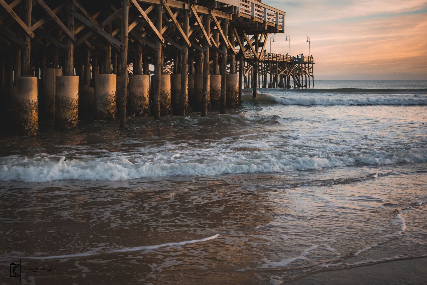 Wooden fishing pier extends over crashing waves at sunset with warm golden light illuminating the ocean shore.