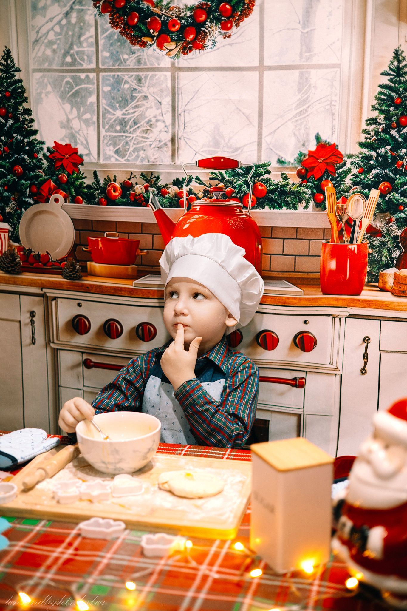 A cozy Christmas kitchen scene with a child in a Santa hat rolling dough on a plaid tablecloth.