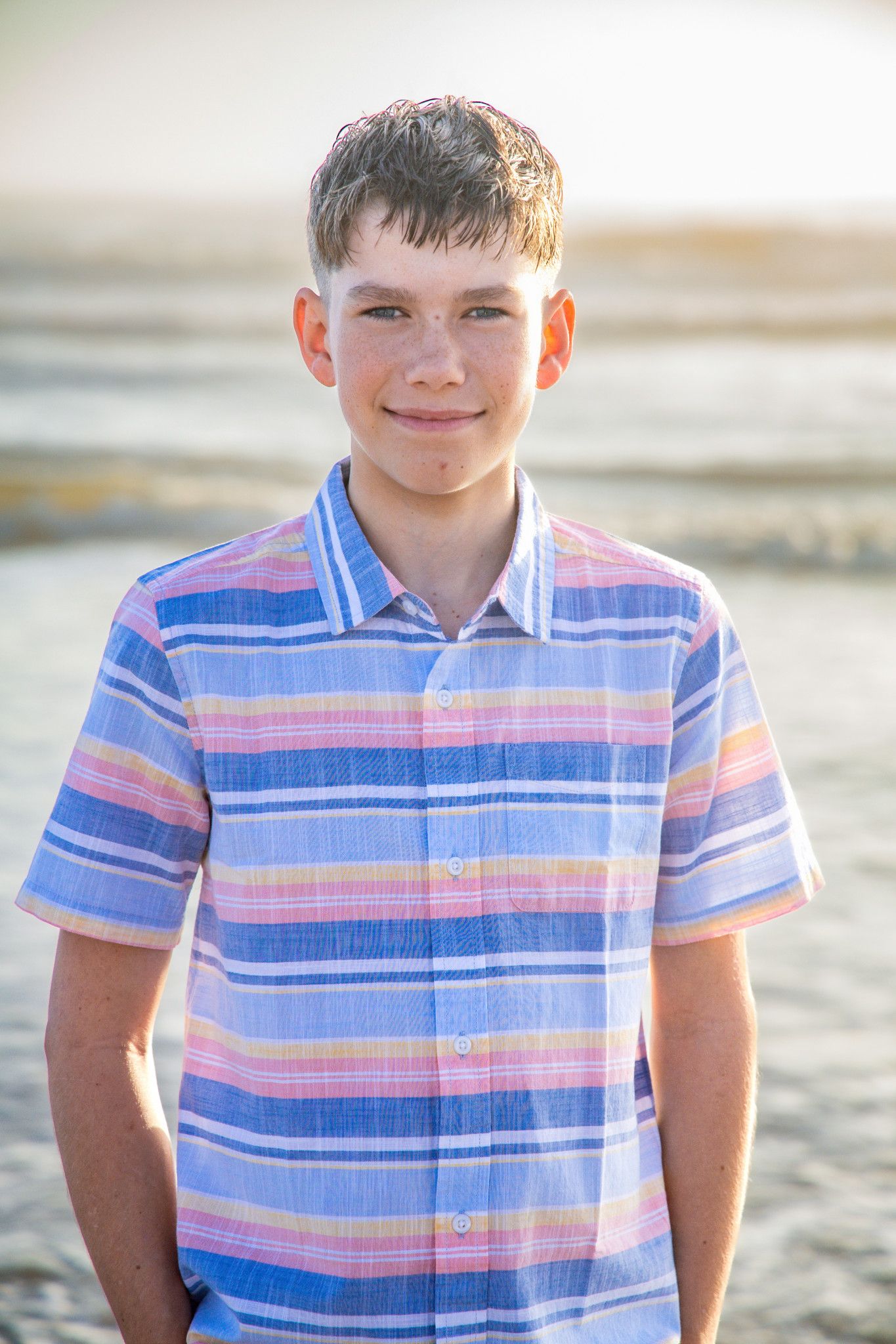 Young person in a striped polo shirt smiles at the camera in a beach setting.