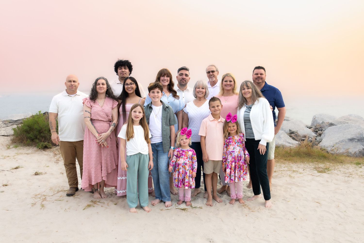 Family group wearing coordinated pink and blue outfits poses together on a sandy beach at sunset.