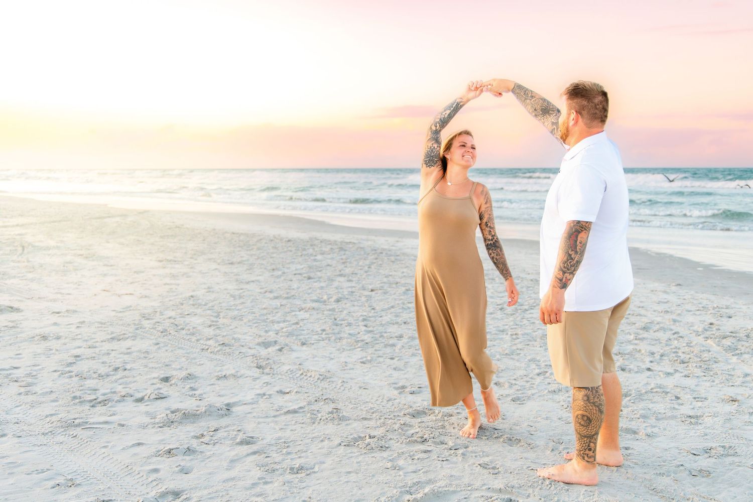 Couple dancing on beach at sunset, man spinning woman in tan dress.