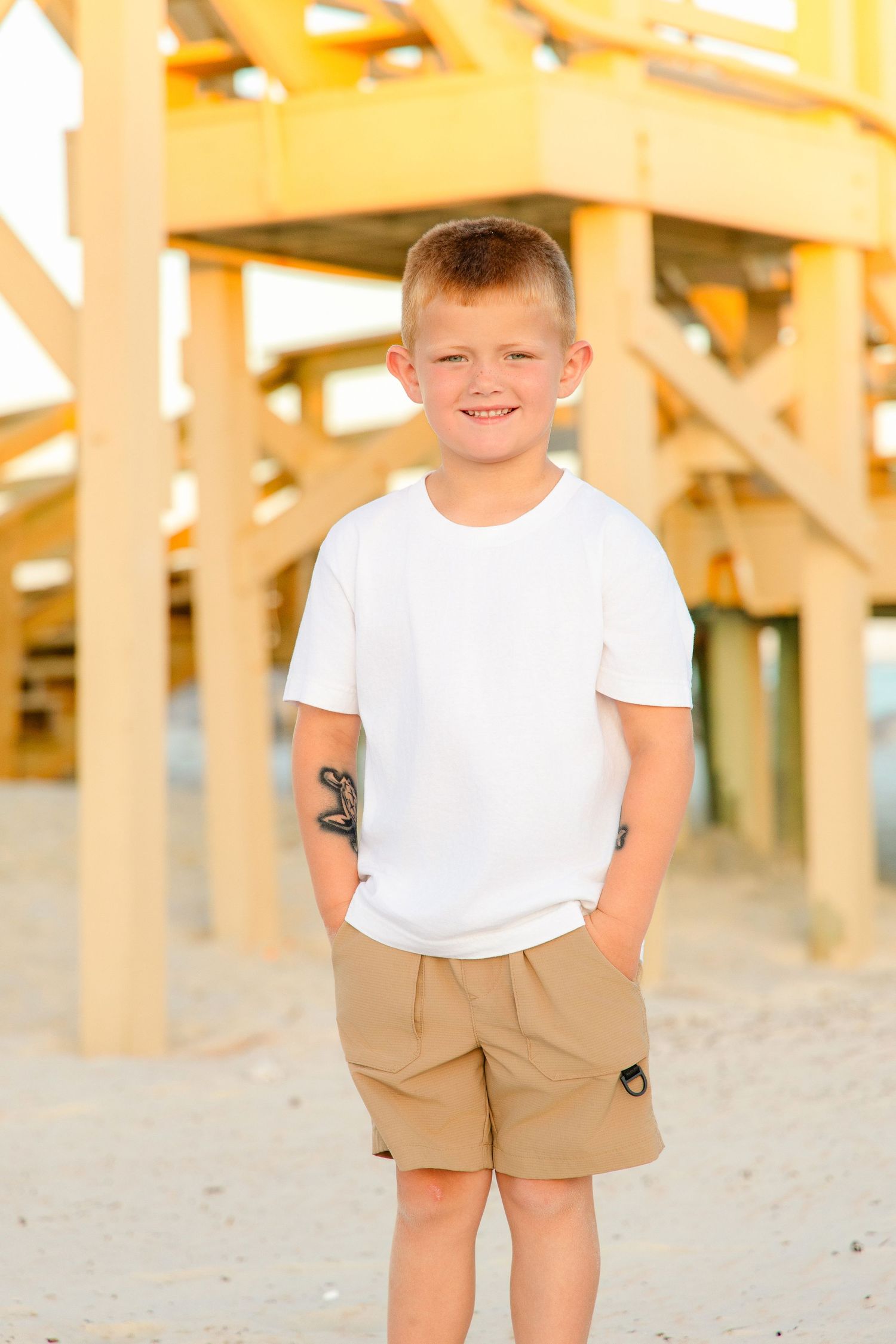 Smiling boy in white t-shirt and khaki shorts standing on sandy beach near yellow wooden pier structure.