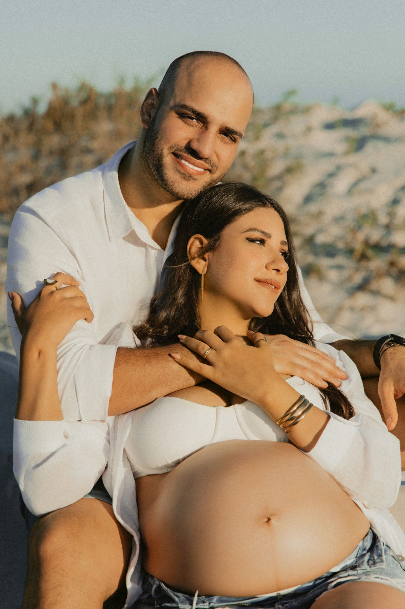 Couple posing for maternity photo shoot on beach, man embracing pregnant woman in white crop top and denim shorts.