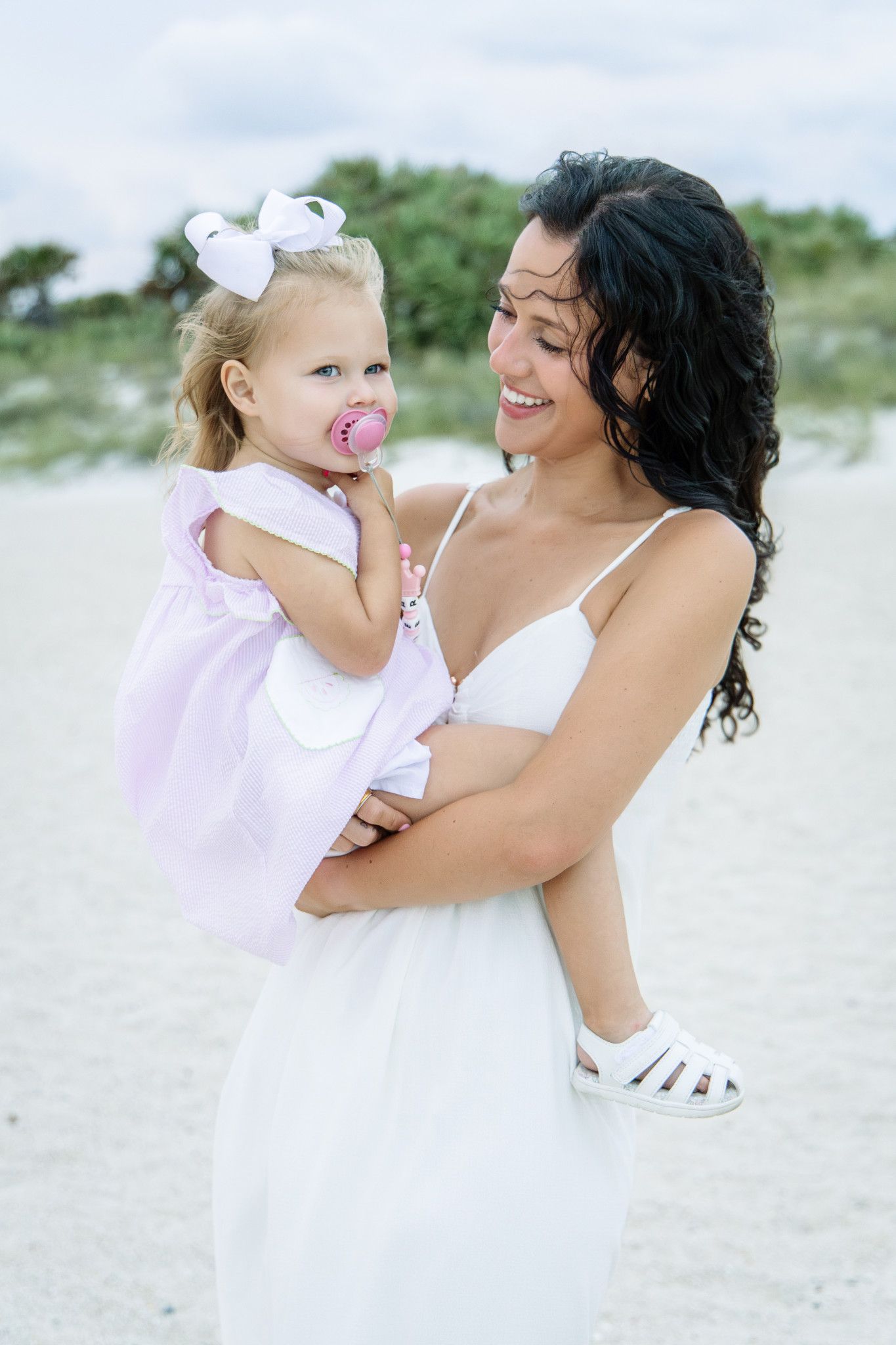 A smiling mother in a white dress holds her young blonde daughter in a pink dress on a sandy beach.