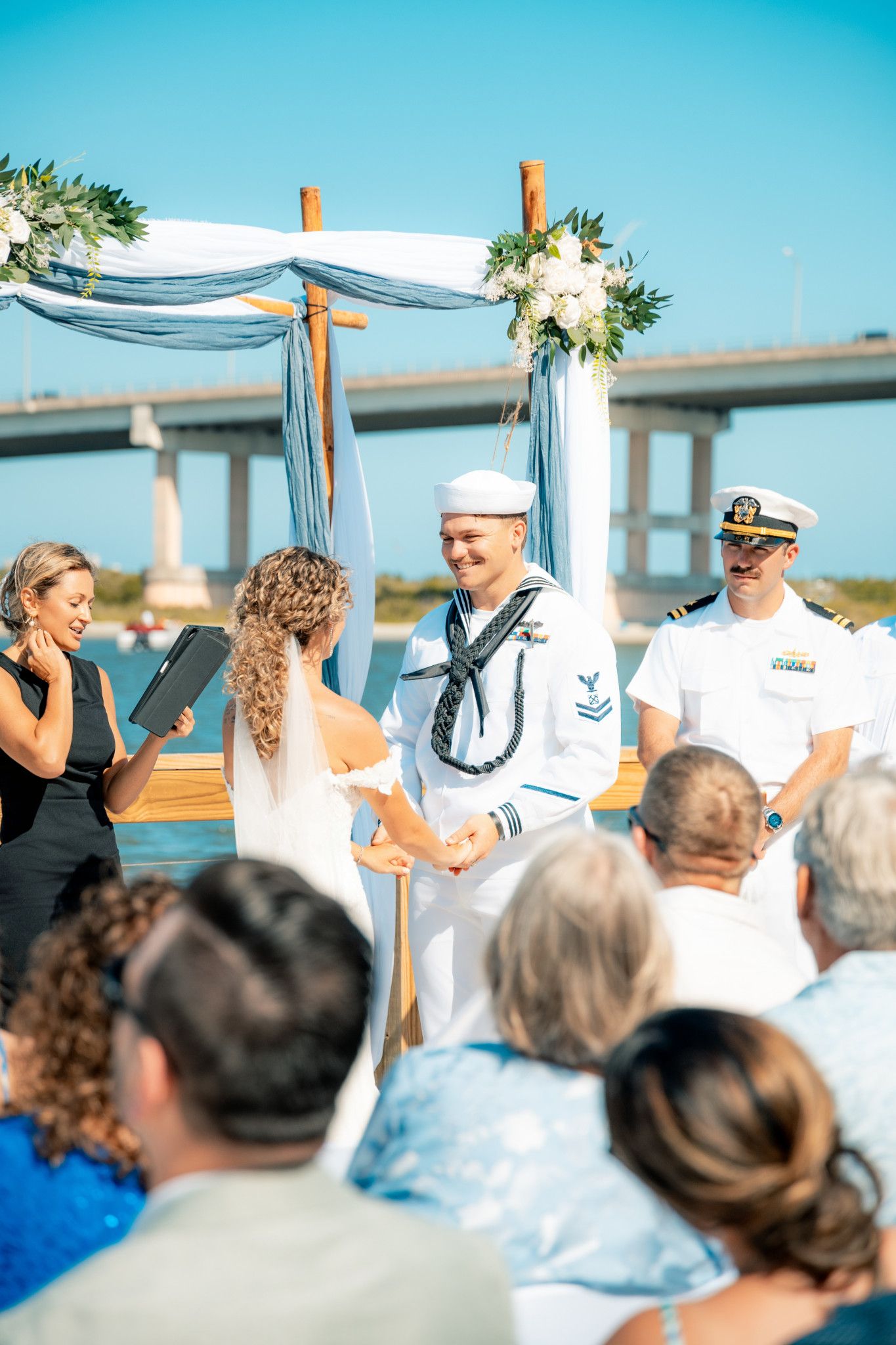 Navy sailor groom smiling at bride during outdoor waterfront wedding ceremony under floral arch with bridge backdrop.