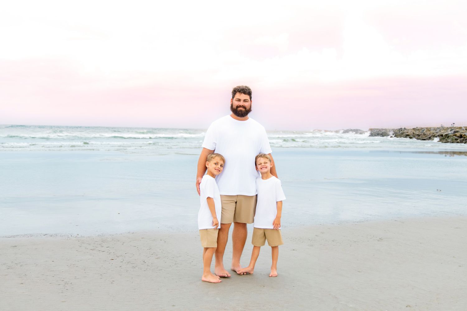A father and two young children in matching white shirts and khaki shorts pose on a sandy beach at sunset.