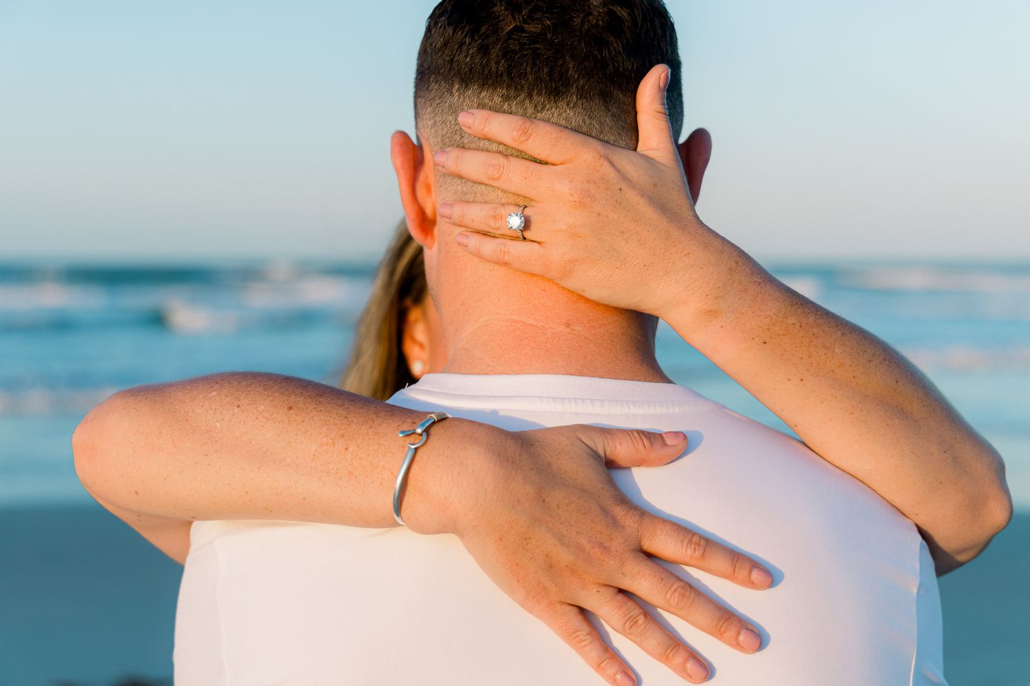 Couple embracing on beach at sunset, woman showing engagement ring while hugging man from behind.